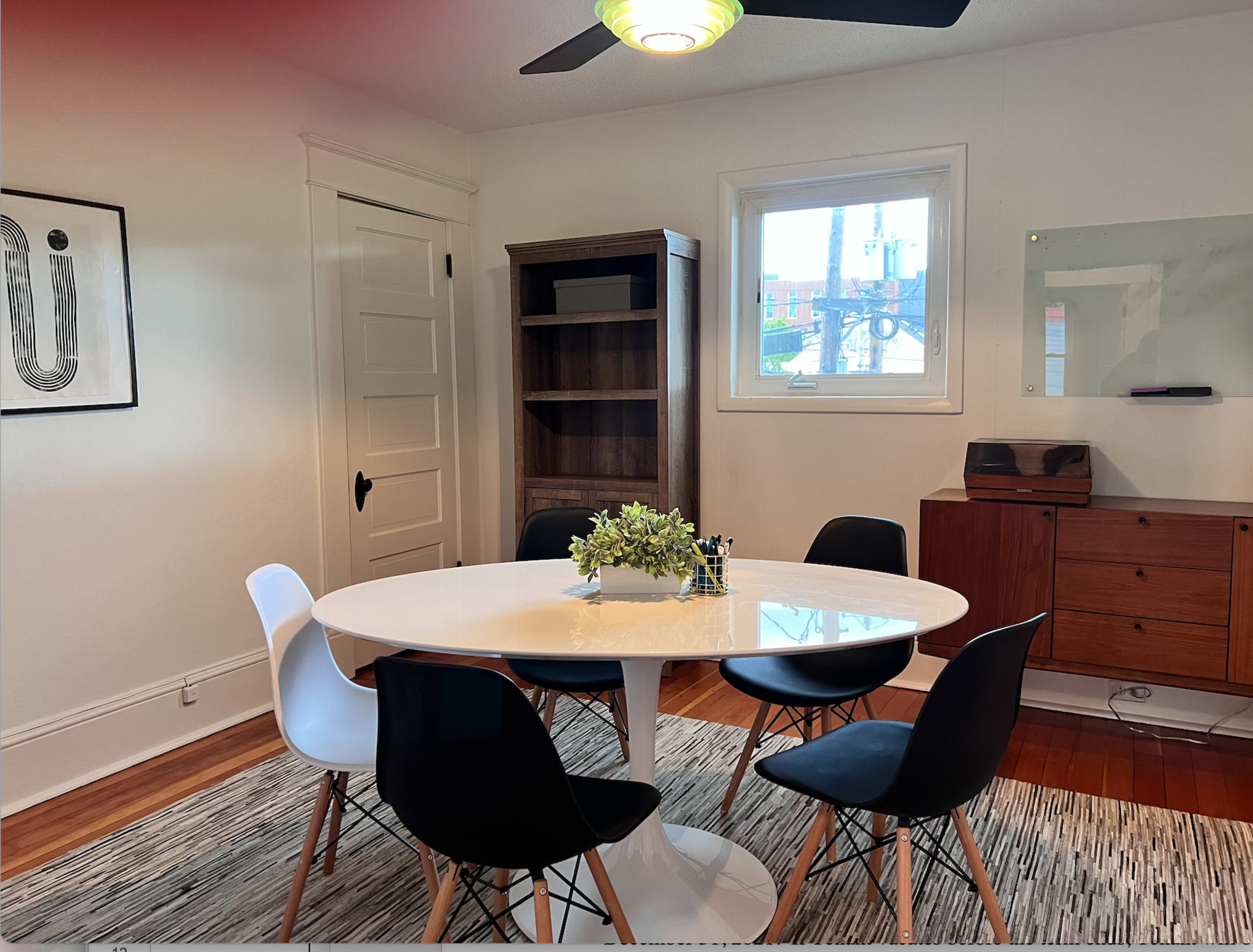 A round white dining table is surrounded by four chairs, with a plant centerpiece, in a well-lit room featuring a bookshelf and a wooden sideboard.