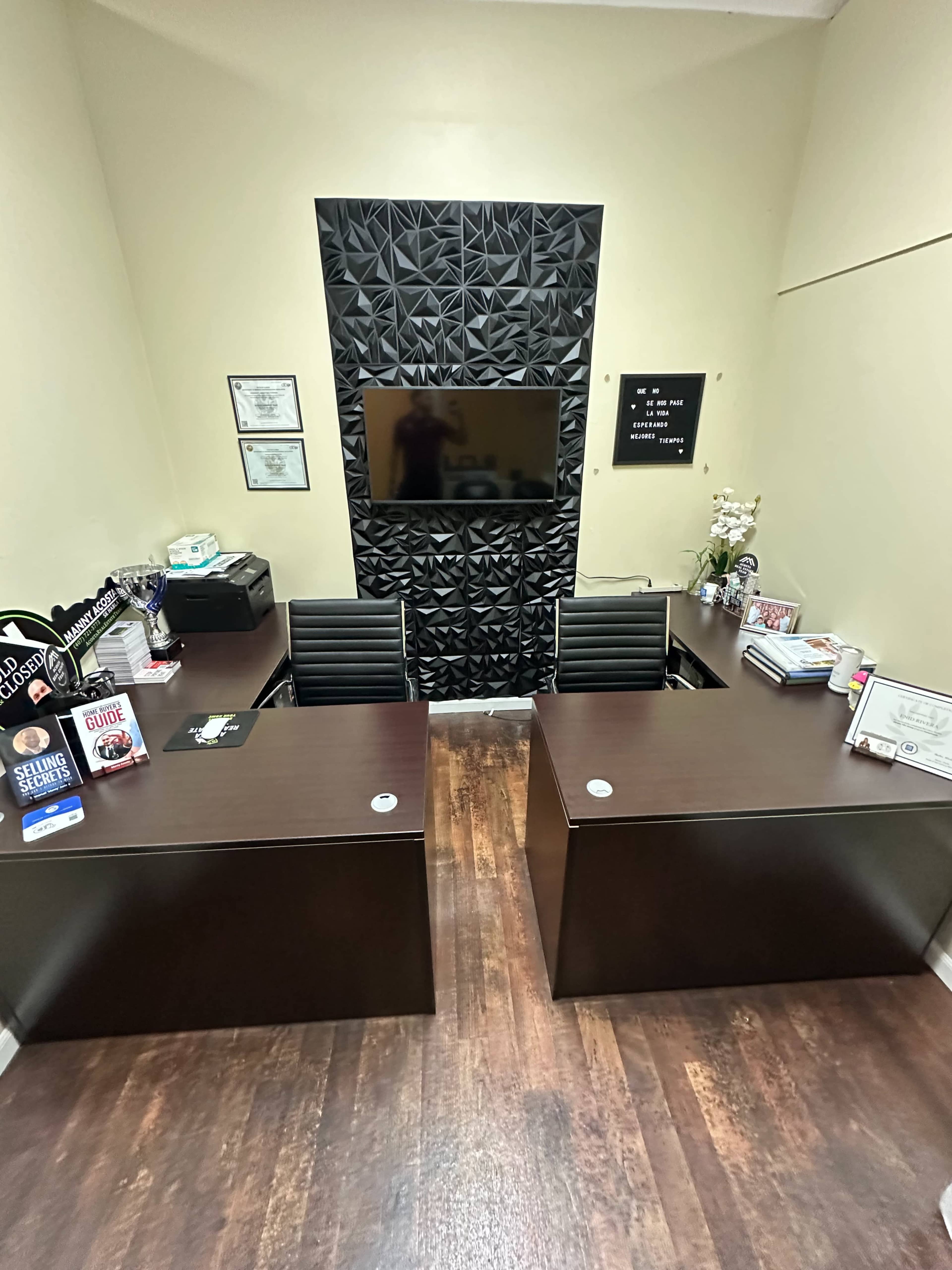 The image shows an office space with two dark wood desks facing each other, a textured black wall with a television mounted on it, and various items and certificates placed on the desks.