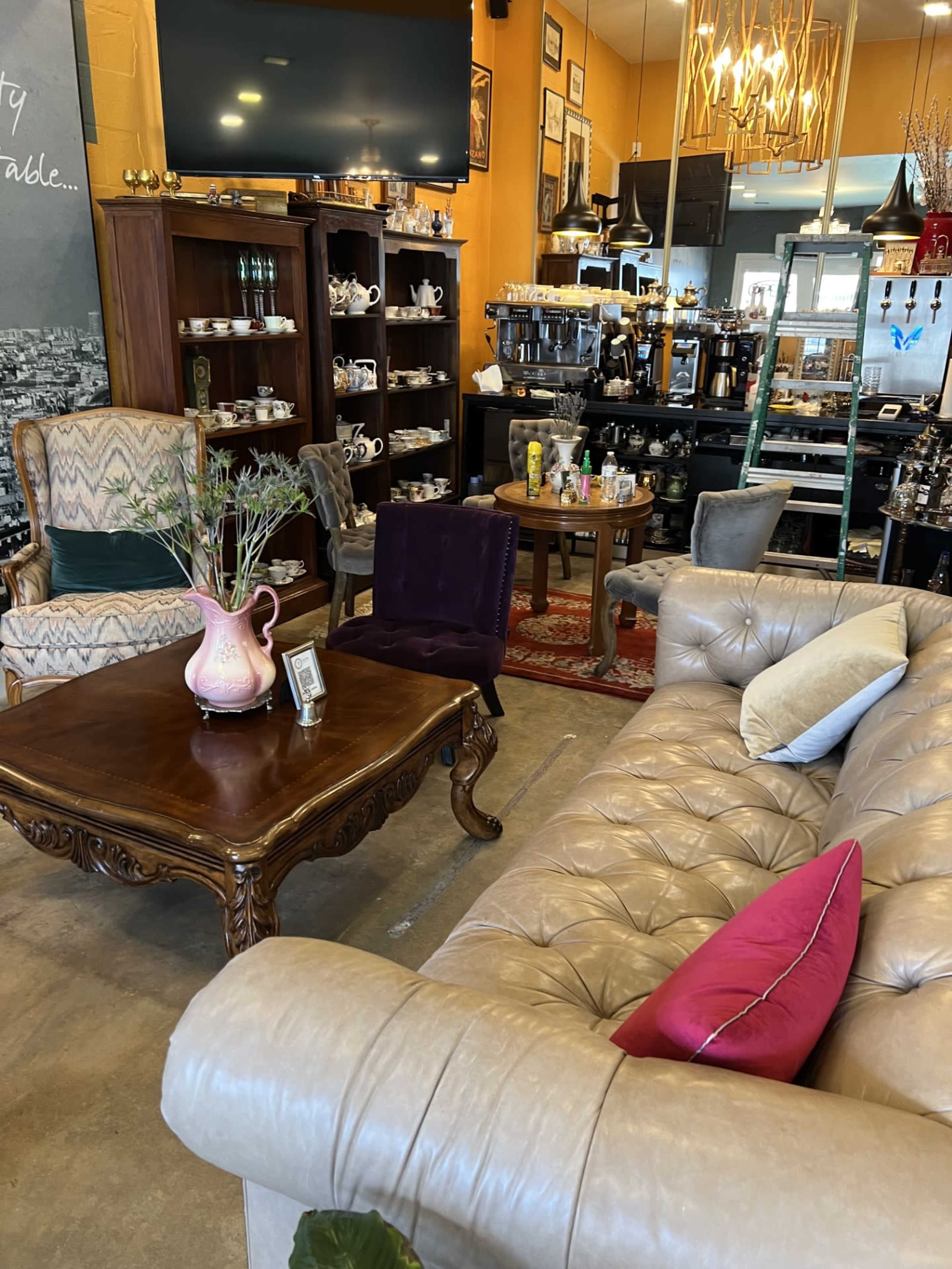 The image shows a cozy café interior with a mix of vintage furniture, including a leather sofa, an ornate wooden coffee table, and shelves filled with coffee cups and decor.