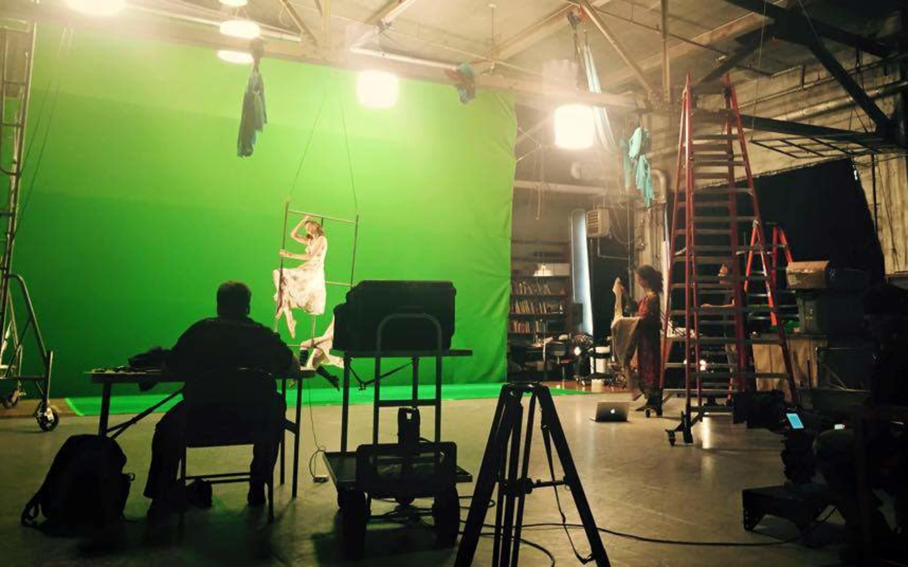 The image shows a film studio set equipped with a green screen, lighting equipment, a ladder, and a figure hanging from a swing in the background.