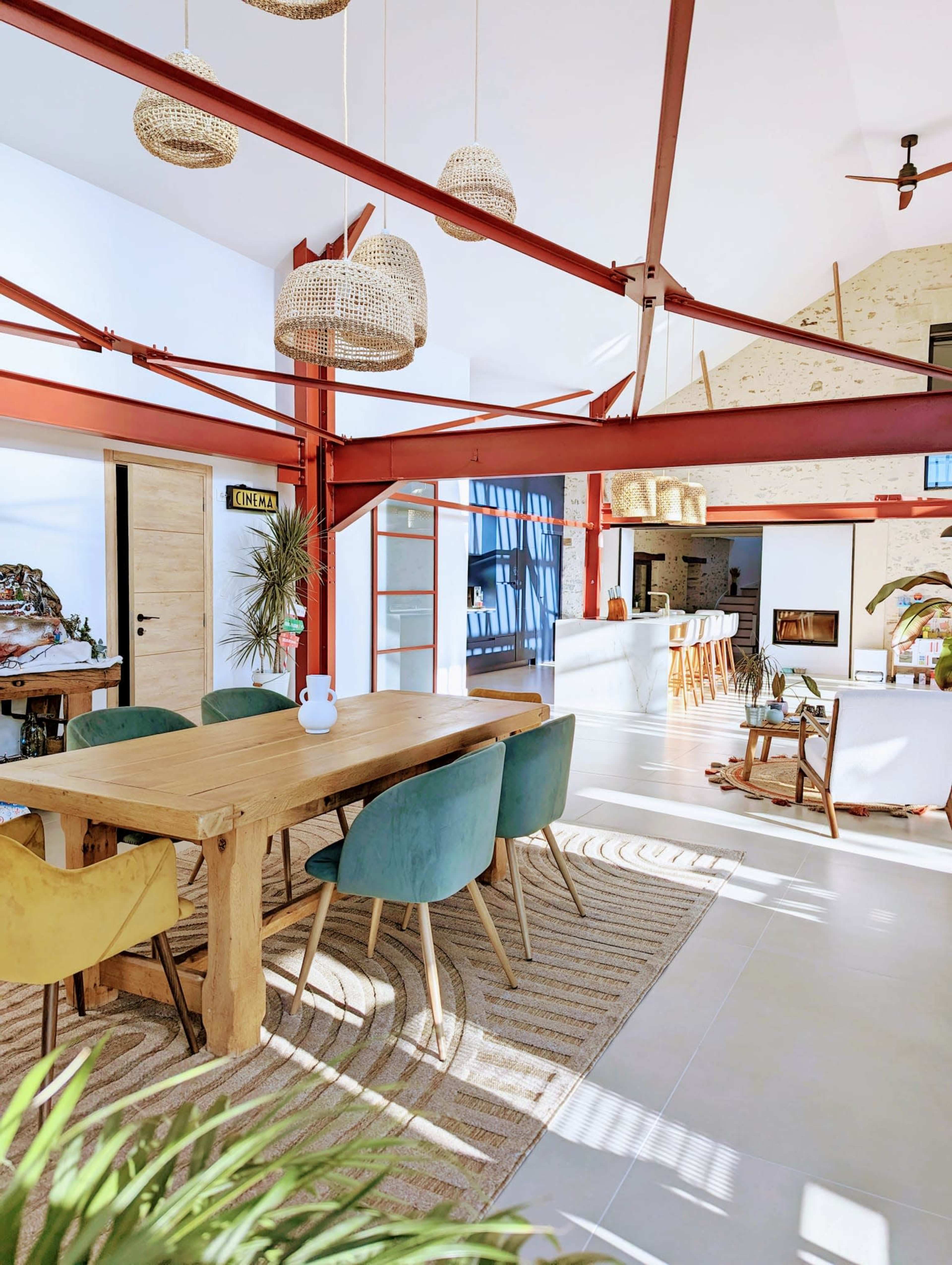 A modern dining area with a wooden table surrounded by colorful chairs, featuring exposed structural beams and pendant lighting.