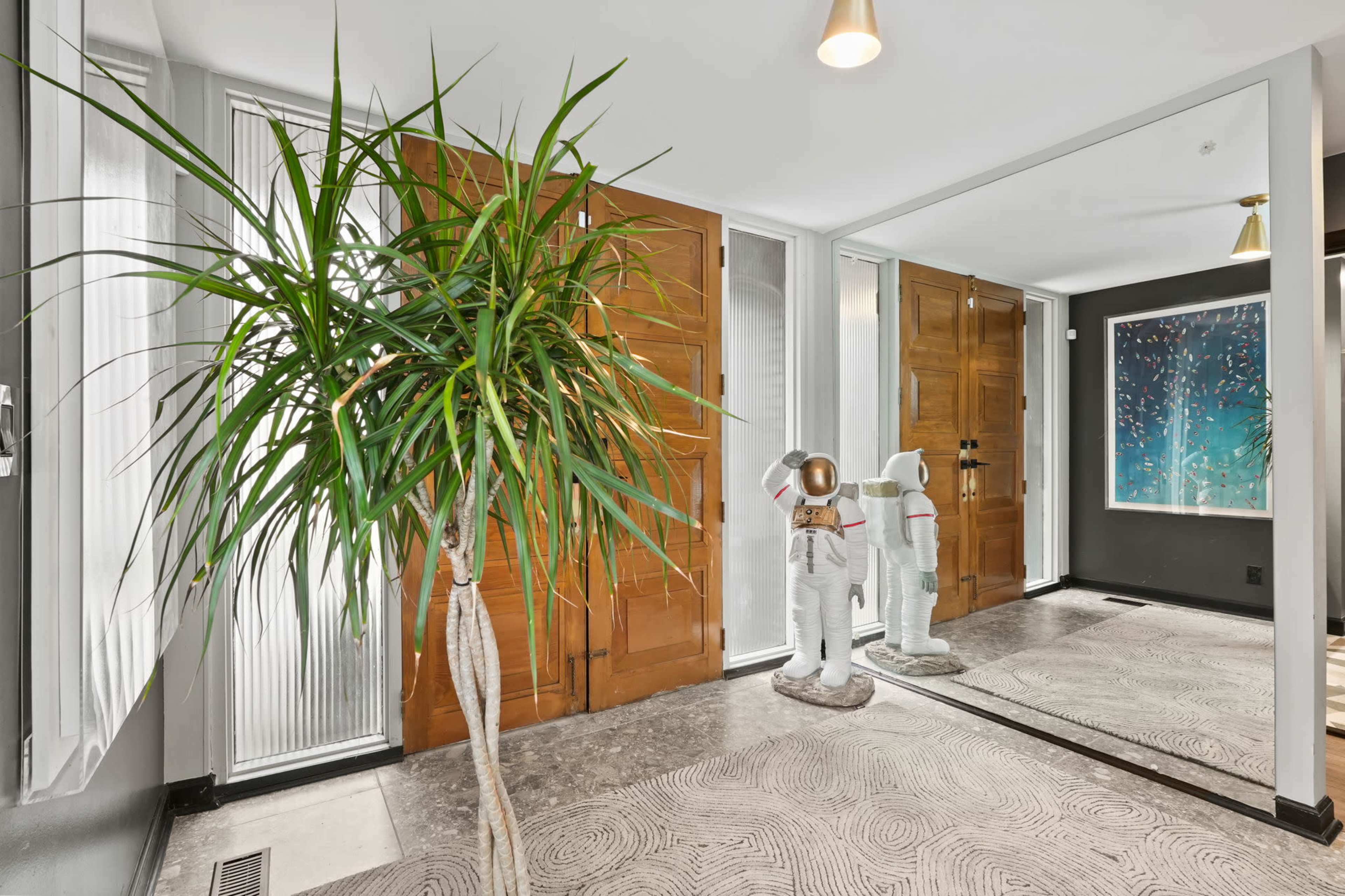 A modern entrance hall with wooden doors, a decorative plant, a space-themed astronaut figure, and a colorful abstract artwork on the wall.