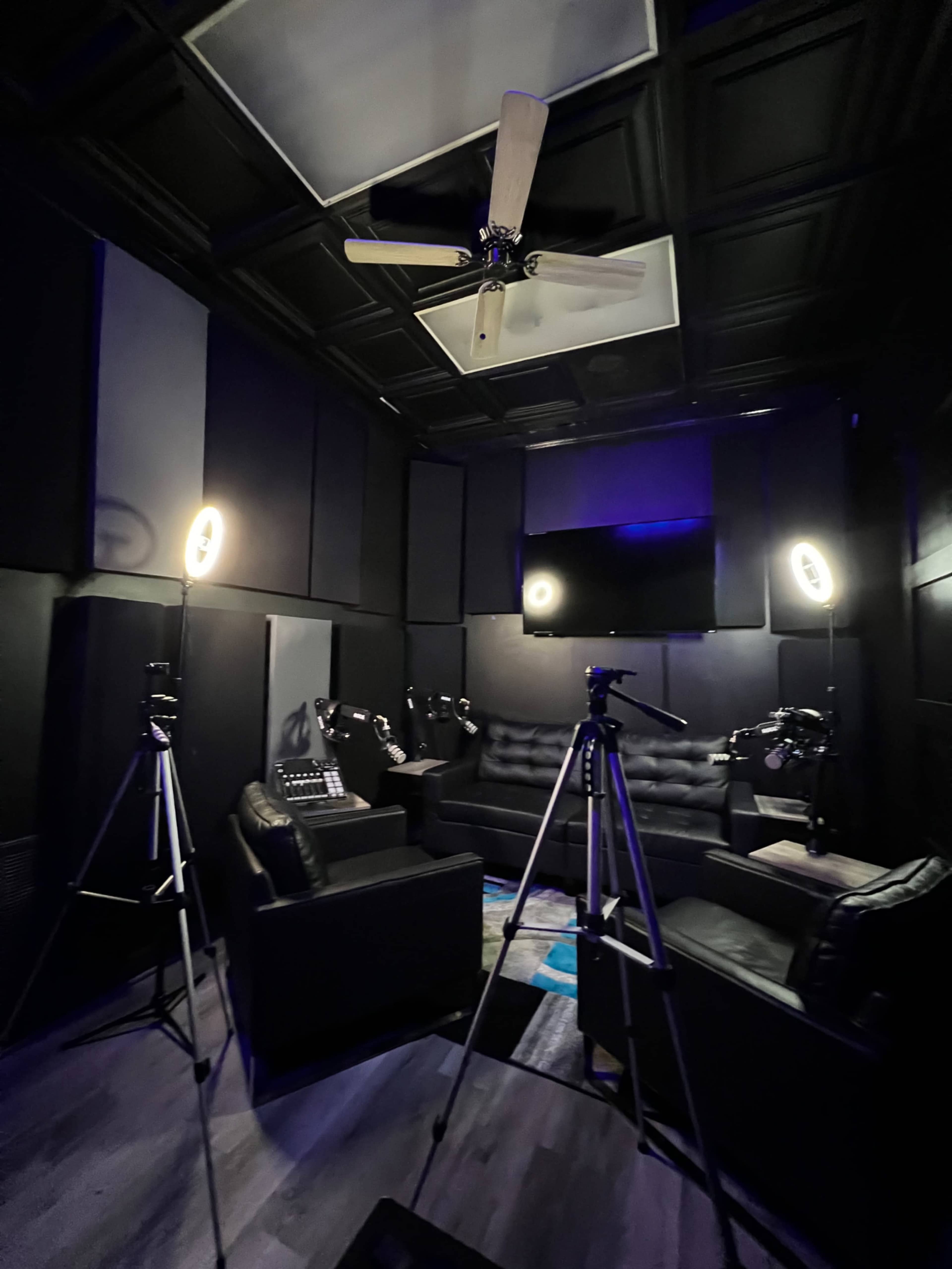 A dimly lit recording studio features black walls, a ceiling fan, a large television on the wall, and three tripods with cameras pointed towards a black leather sofa set.