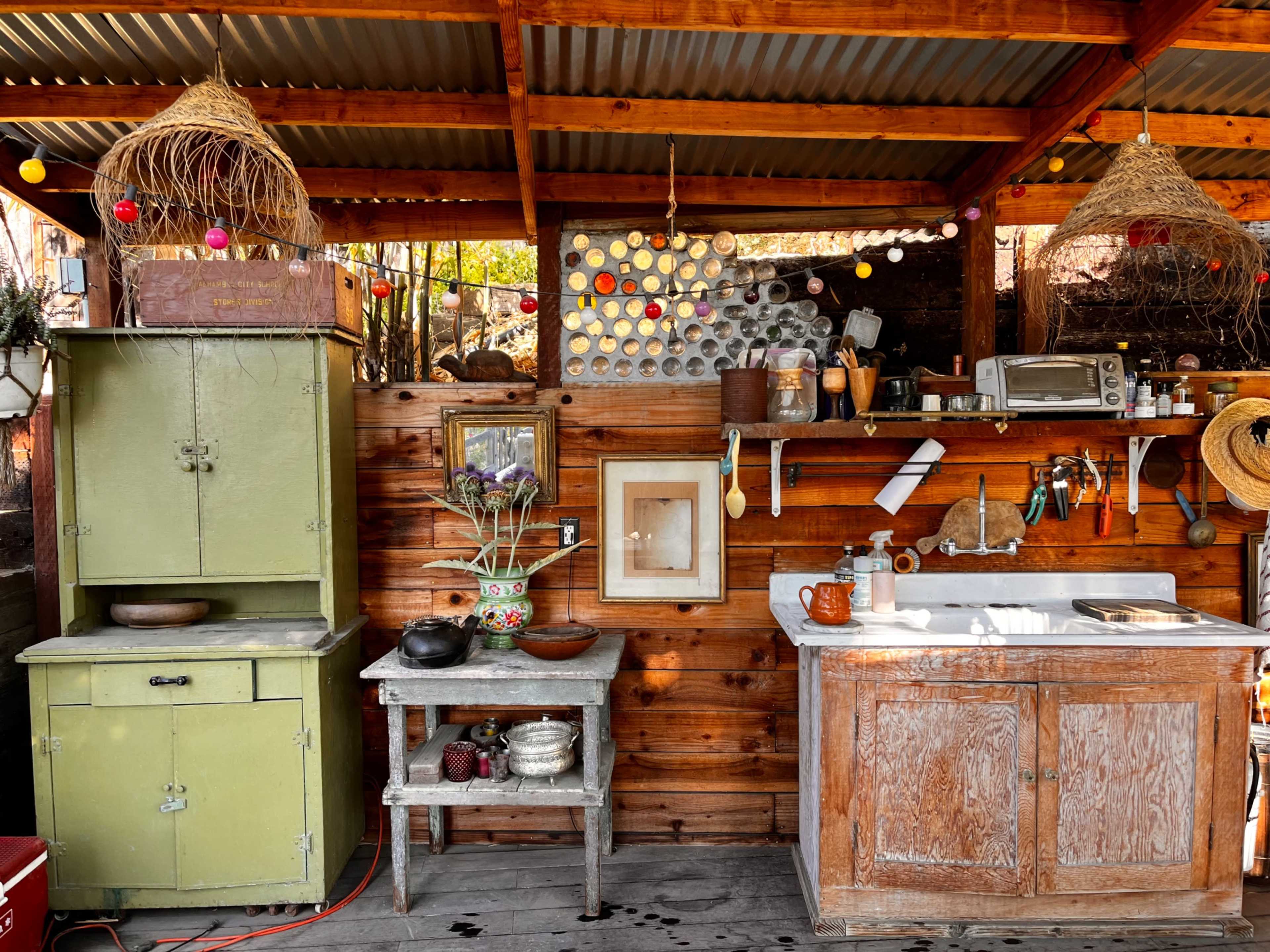 The image shows a rustic wooden kitchen space with a sink, a green cupboard, and a small table, adorned with decorative items and hanging light fixtures.
