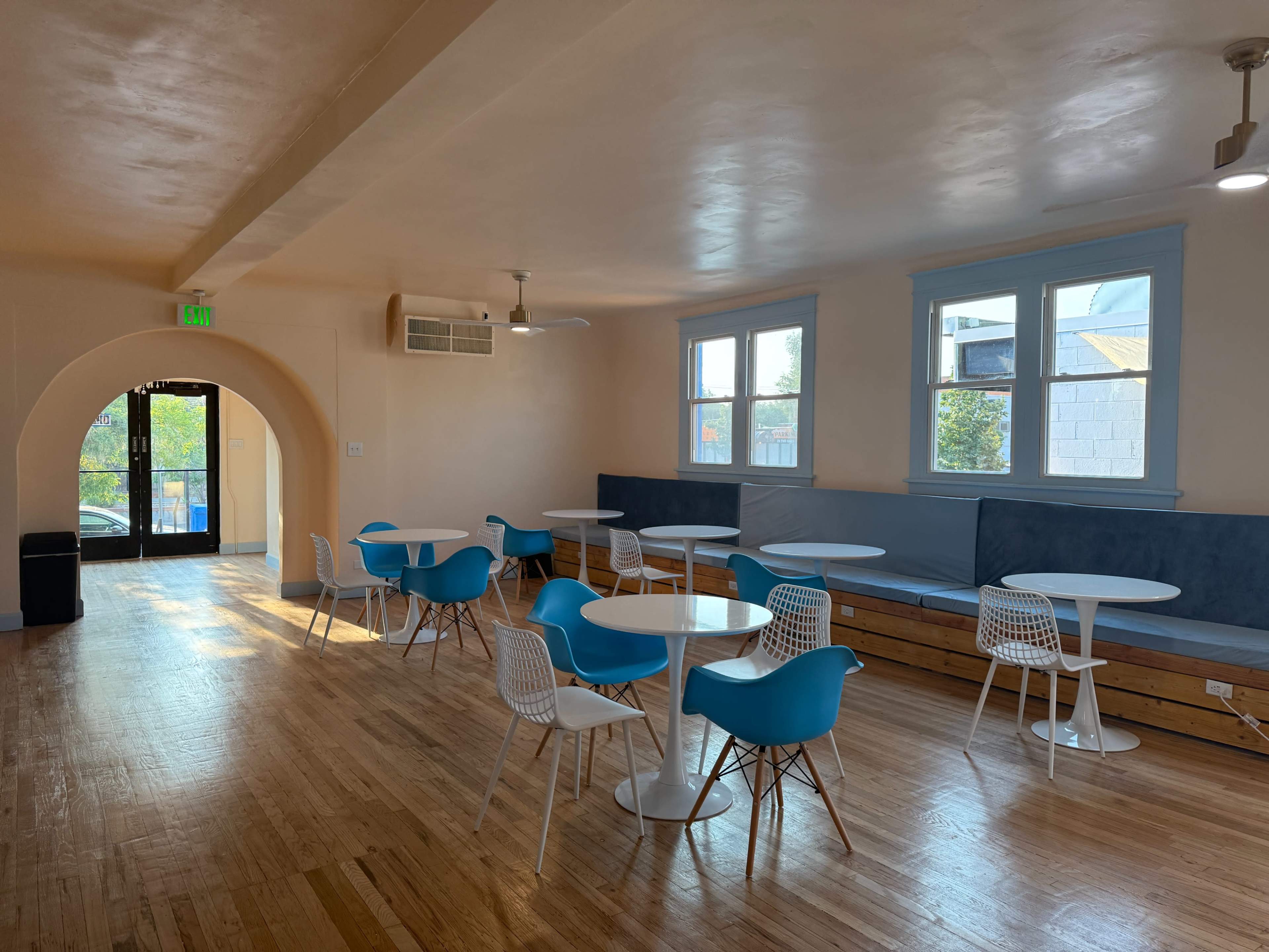 The image shows a spacious, brightly lit room with several blue and white chairs and tables, large windows, and an arched entrance leading outside.