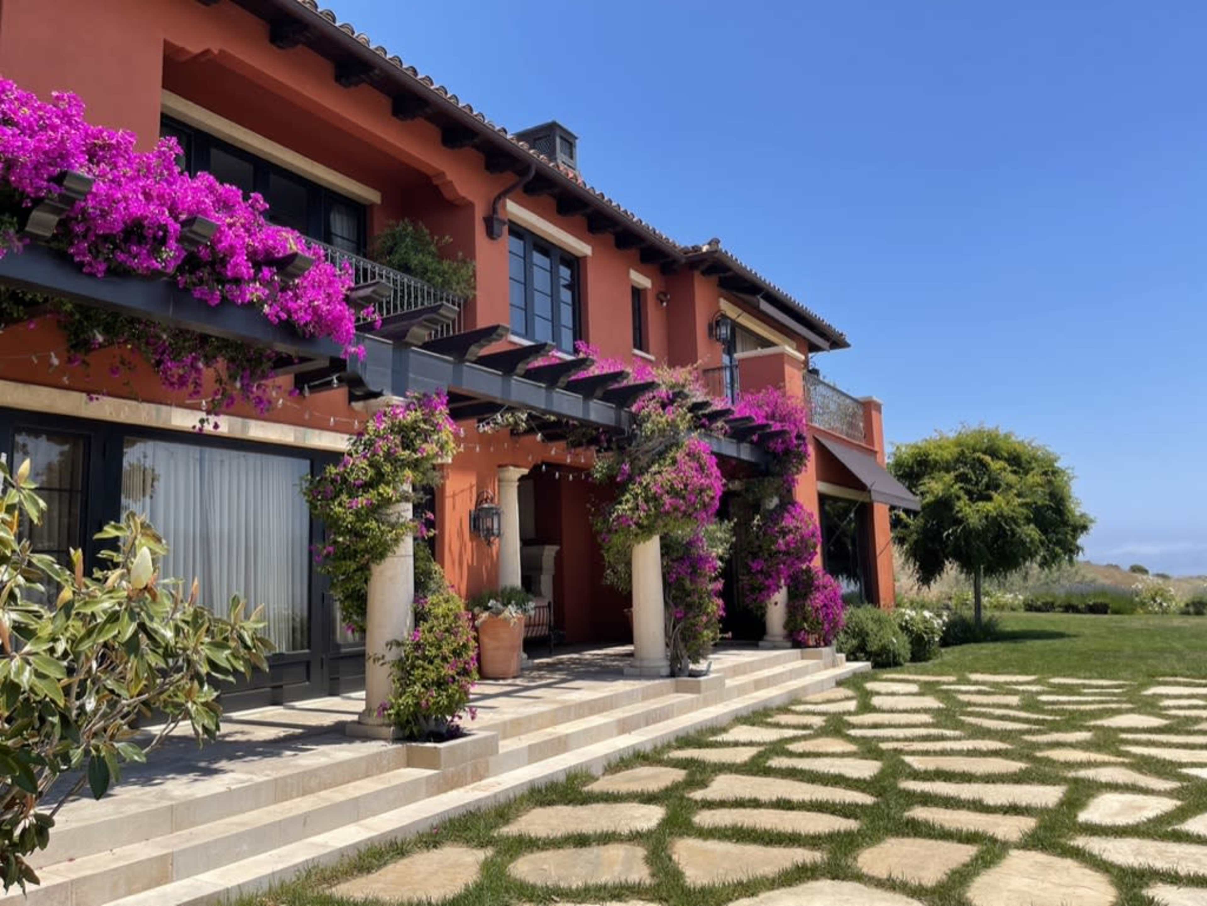 A two-story house with vibrant flower arrangements adorning the exterior, surrounded by a stone pathway and grassy areas.