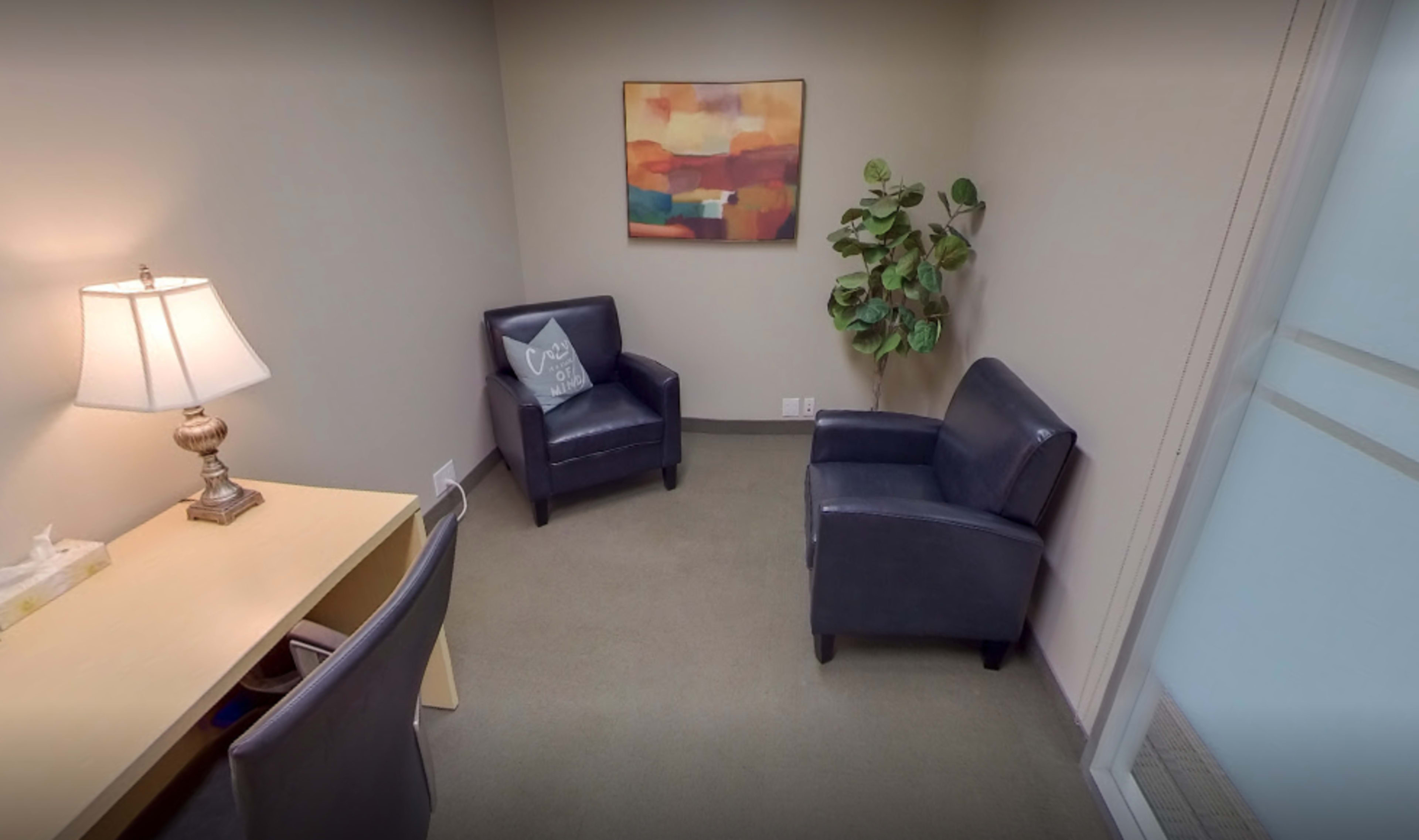 A small office space featuring two leather chairs, a desk with a lamp, and a potted plant, with abstract artwork on the wall.