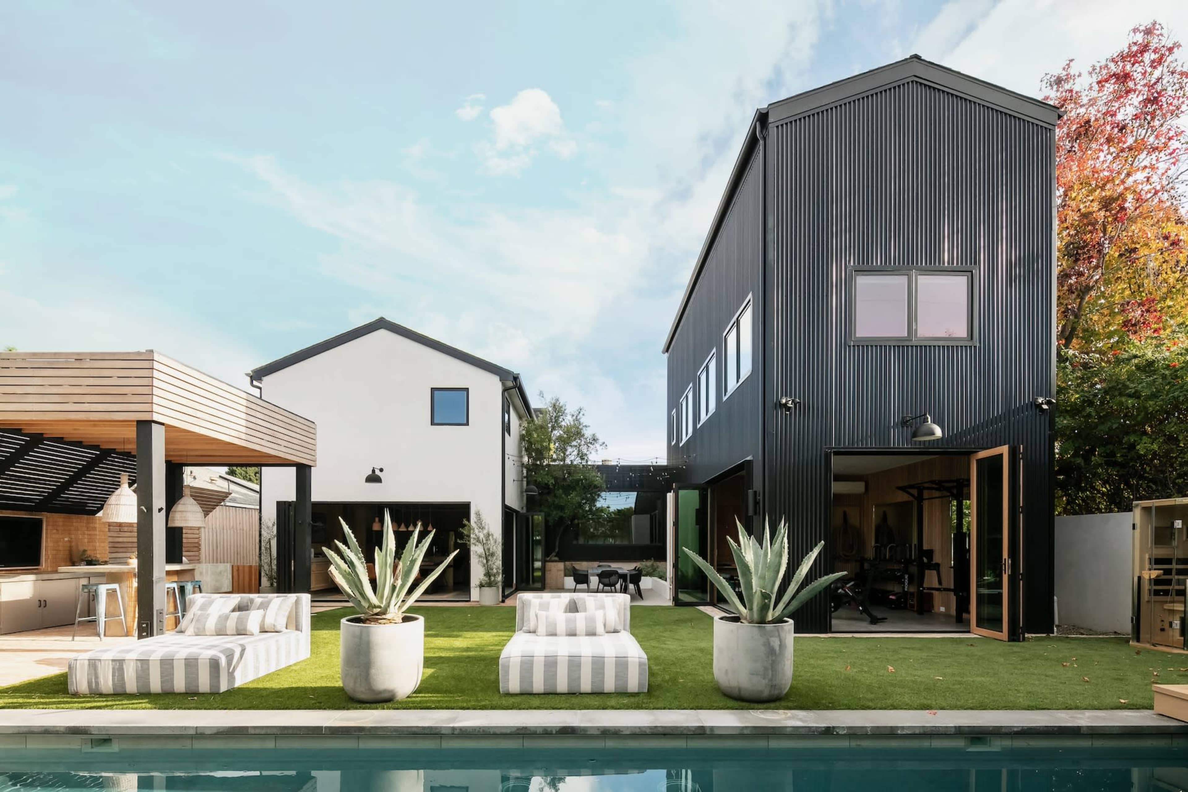 A modern house with a black corrugated metal exterior, accompanied by a white house, a pool, and landscaped seating areas with large planters.