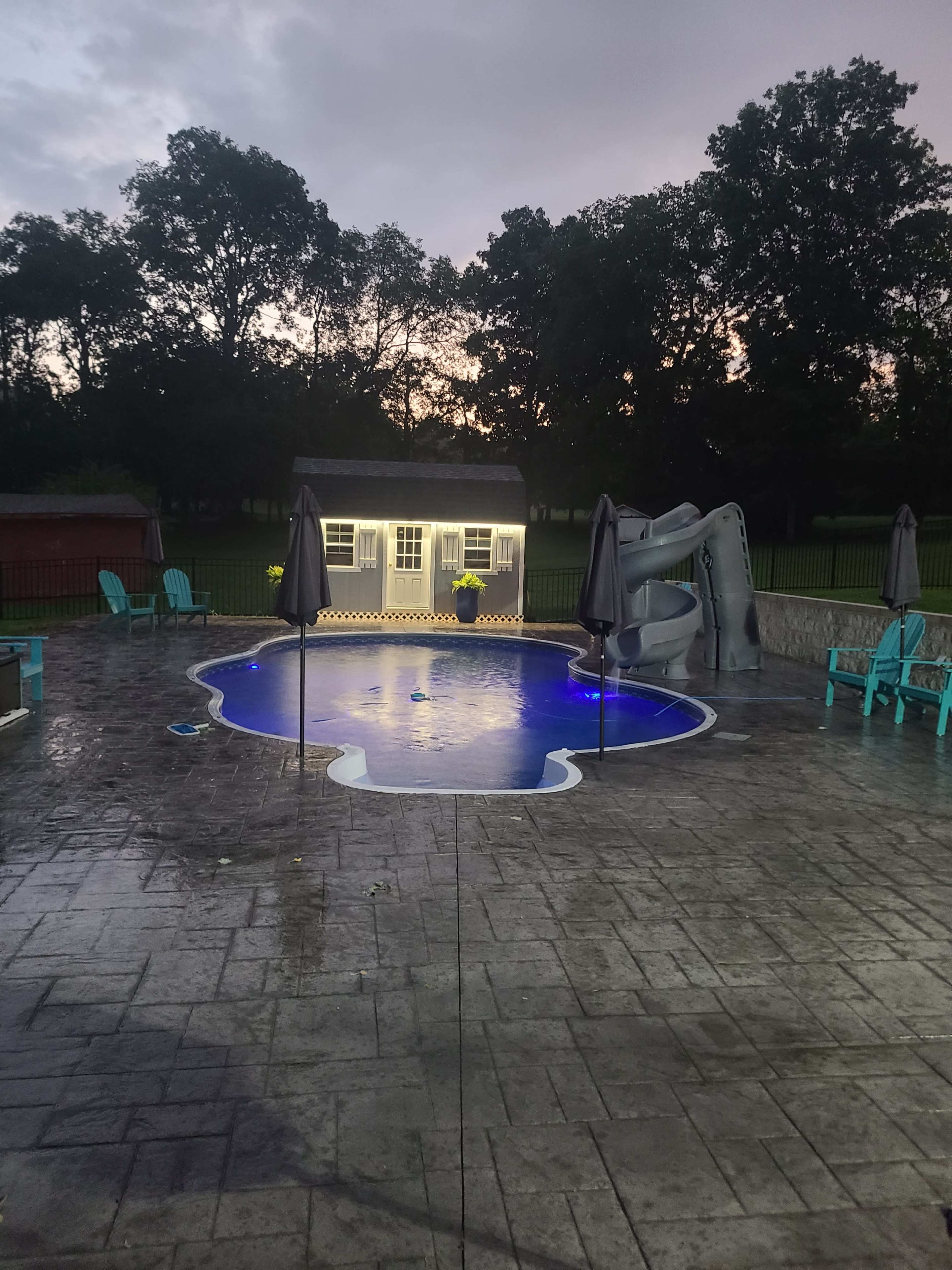 The image shows a residential backyard pool with a slide, surrounded by empty chairs, under an overcast sky during dusk.
