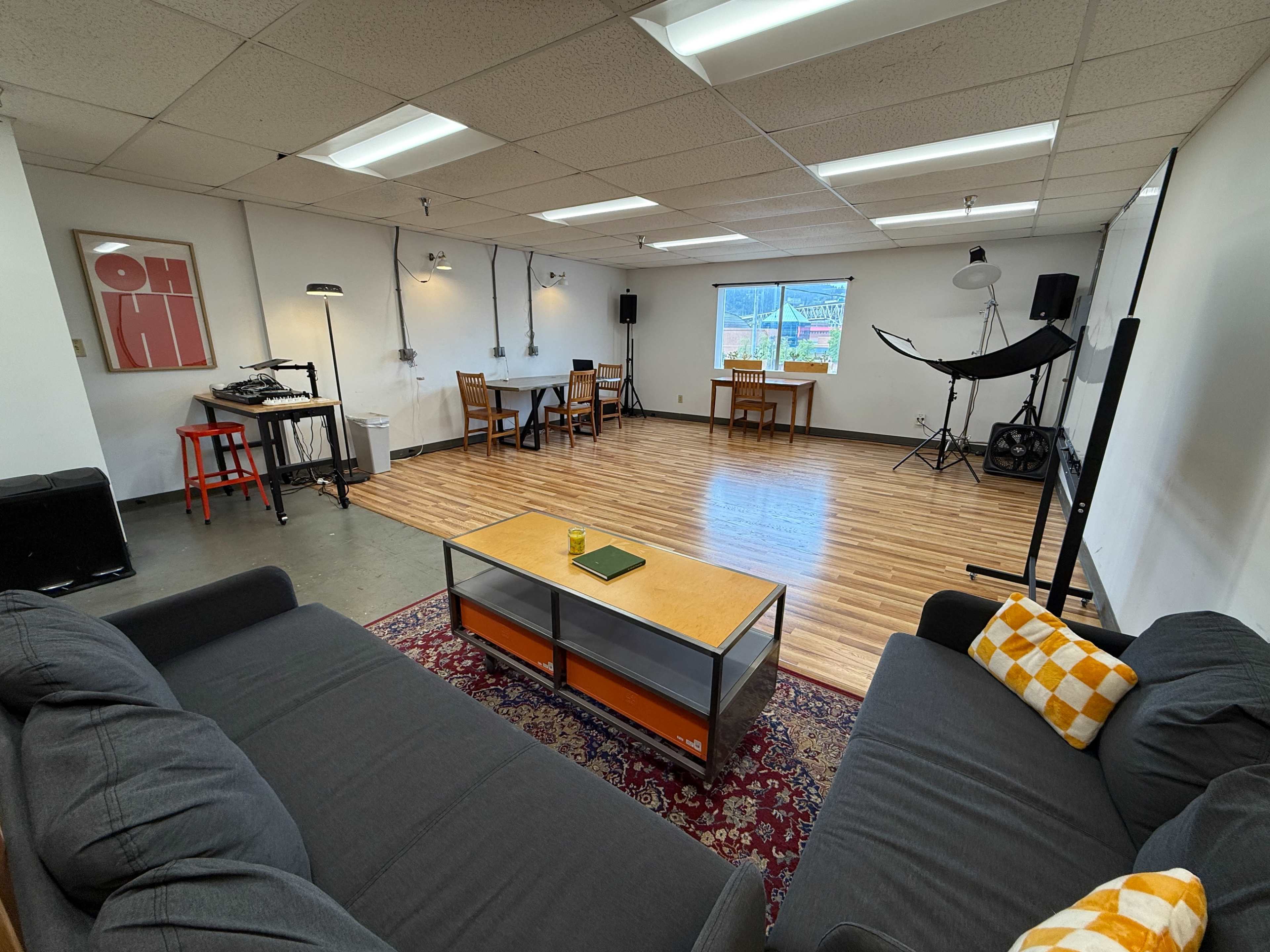 The image shows a spacious room with hardwood flooring, featuring a seating area with two sofas, a coffee table, a dining area with chairs, and equipment including lighting and a backdrop for photography.