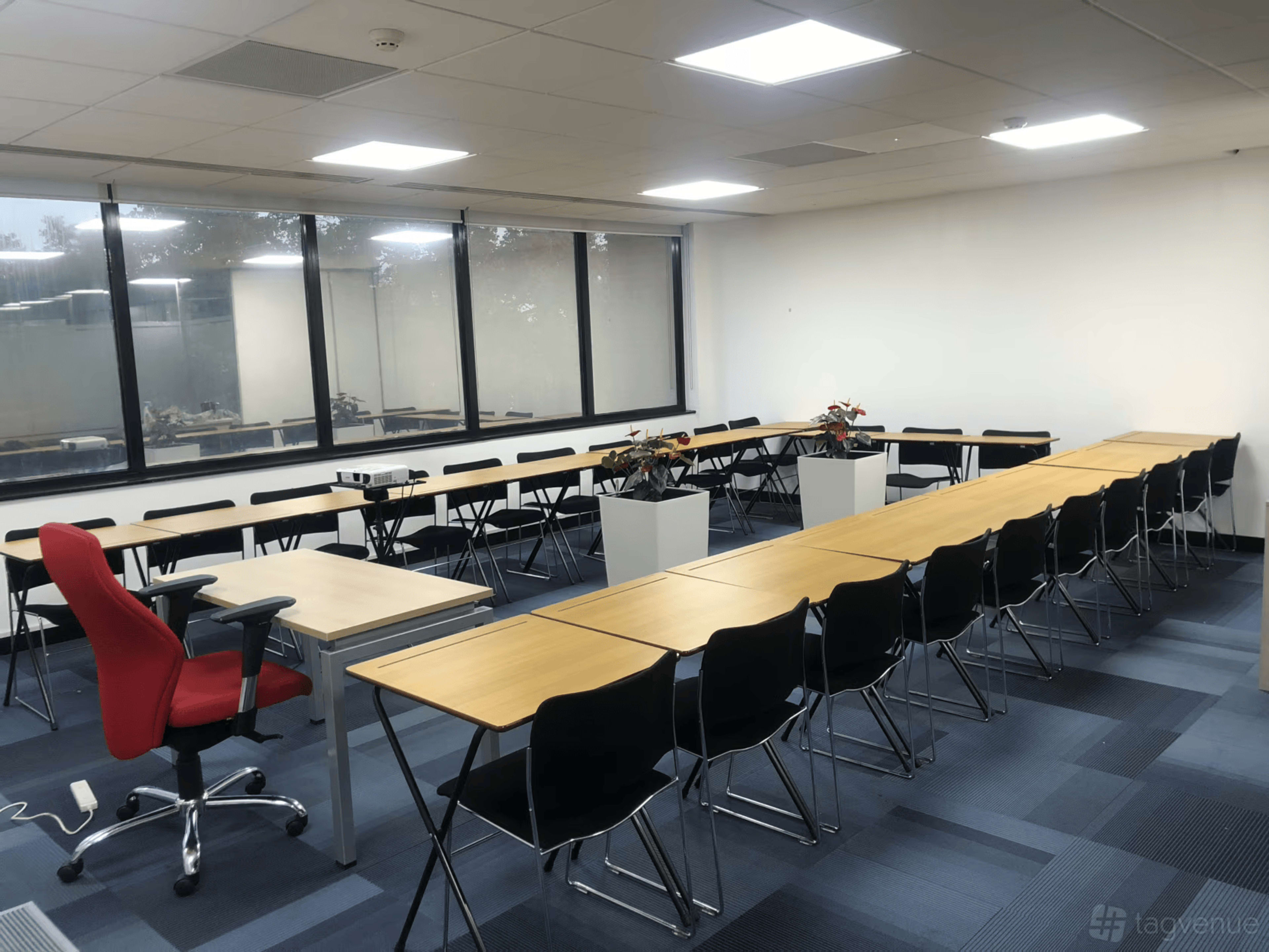 The image shows a conference room with several rows of black chairs and wooden tables, featuring a red chair at the front and two planters with plants.