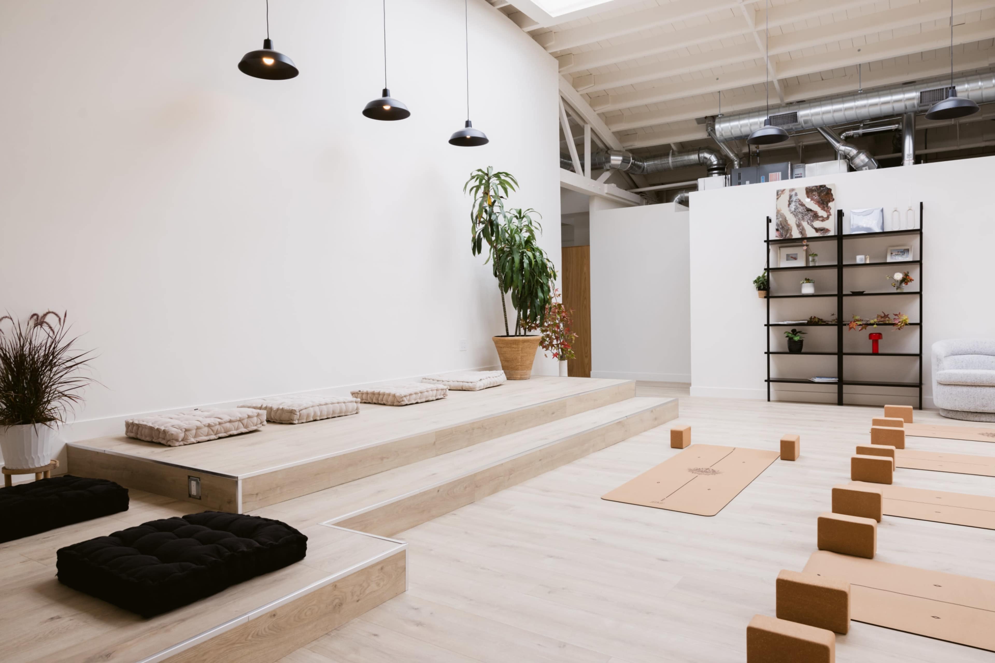 The image shows a minimalist yoga studio with wooden flooring, padded mats arranged on the floor, and stepped seating areas alongside potted plants and simple decor.