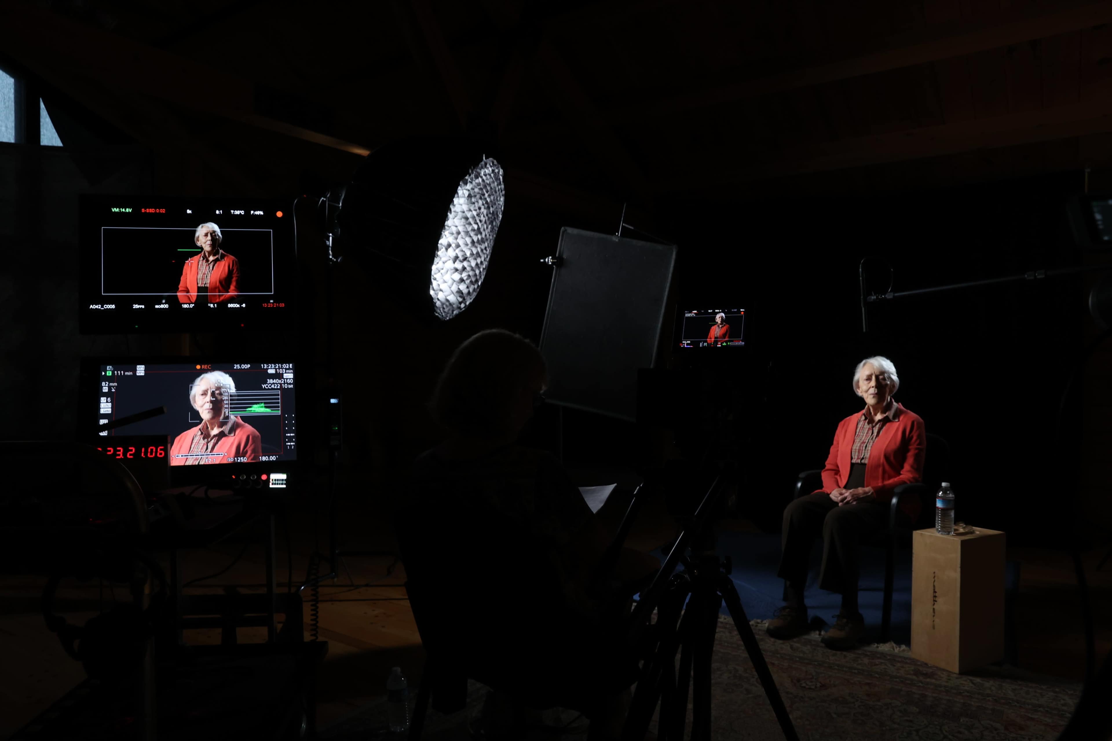 An elderly woman wearing a red jacket is sitting on a wooden box in a lit interview space, surrounded by multiple cameras and lighting equipment.