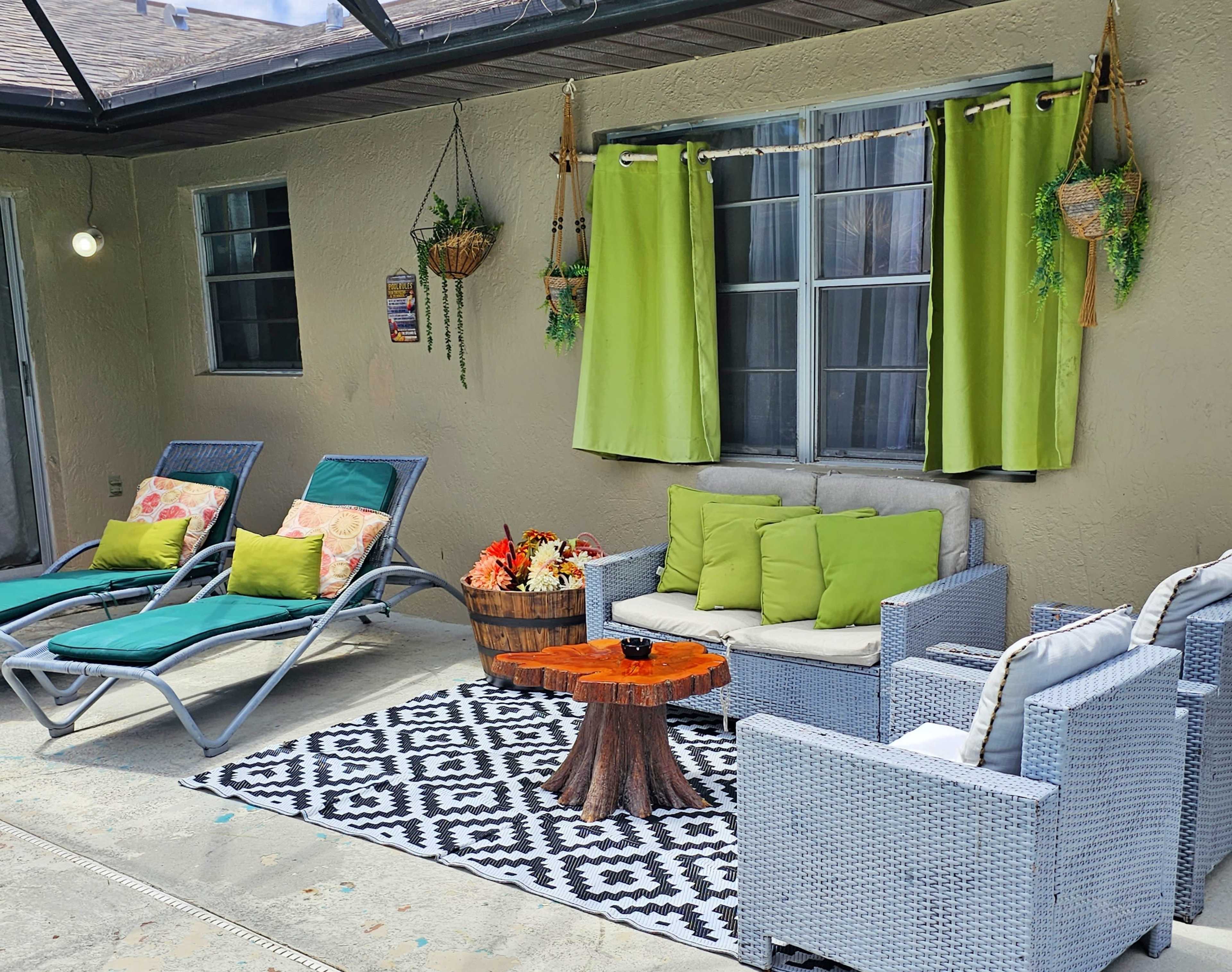 A patio area featuring two lounge chairs, a seating arrangement with green cushions, a wooden side table, decorative curtains, and plants hanging on the walls.