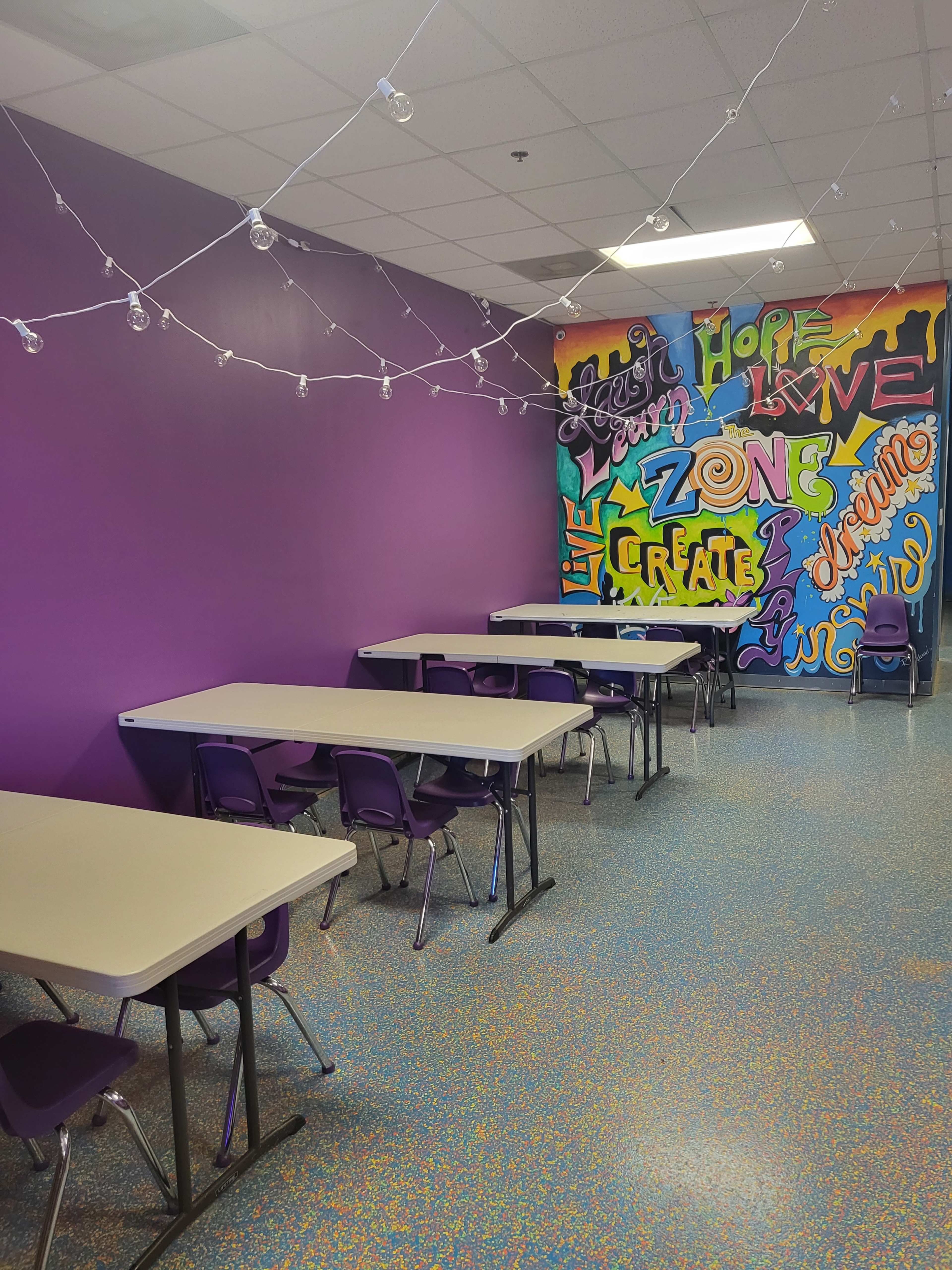 A brightly colored room with several tables and chairs arranged neatly, featuring a vibrant mural on one wall.