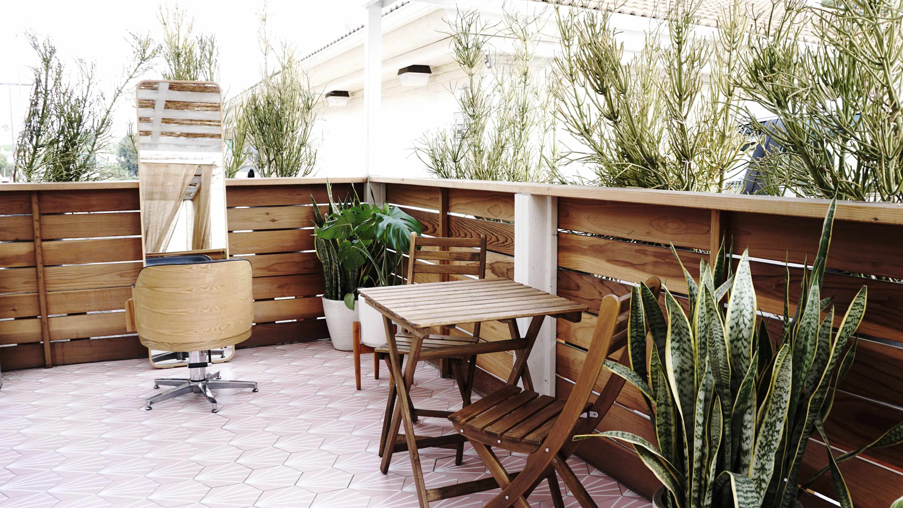 A wooden balcony features a small table and chairs surrounded by tall plants and a mirror.