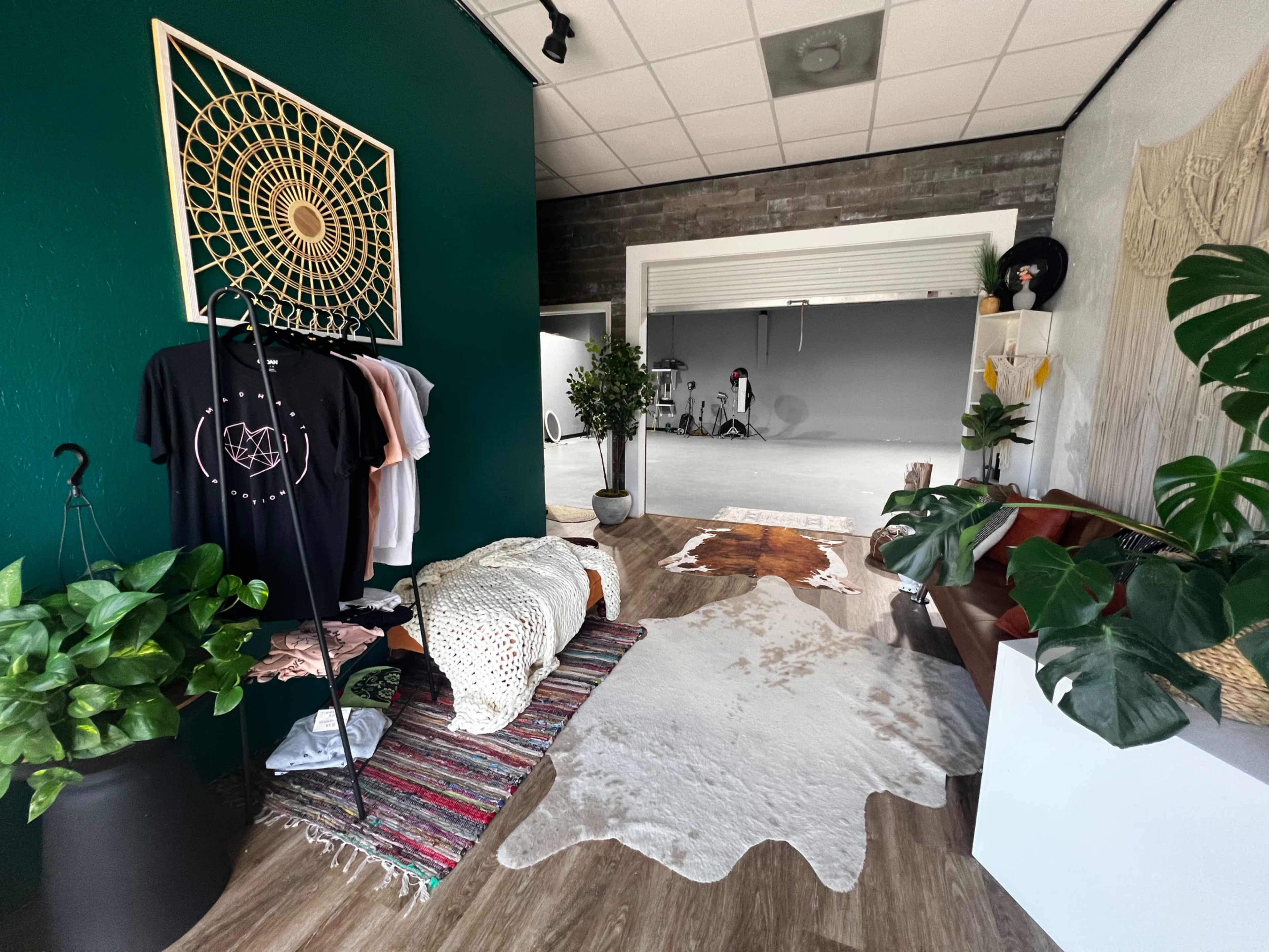 A boutique interior features a green accent wall, a macramé wall hanging, clothing displays, a cowhide rug, potted plants, and a seating area, with an open space in the background.