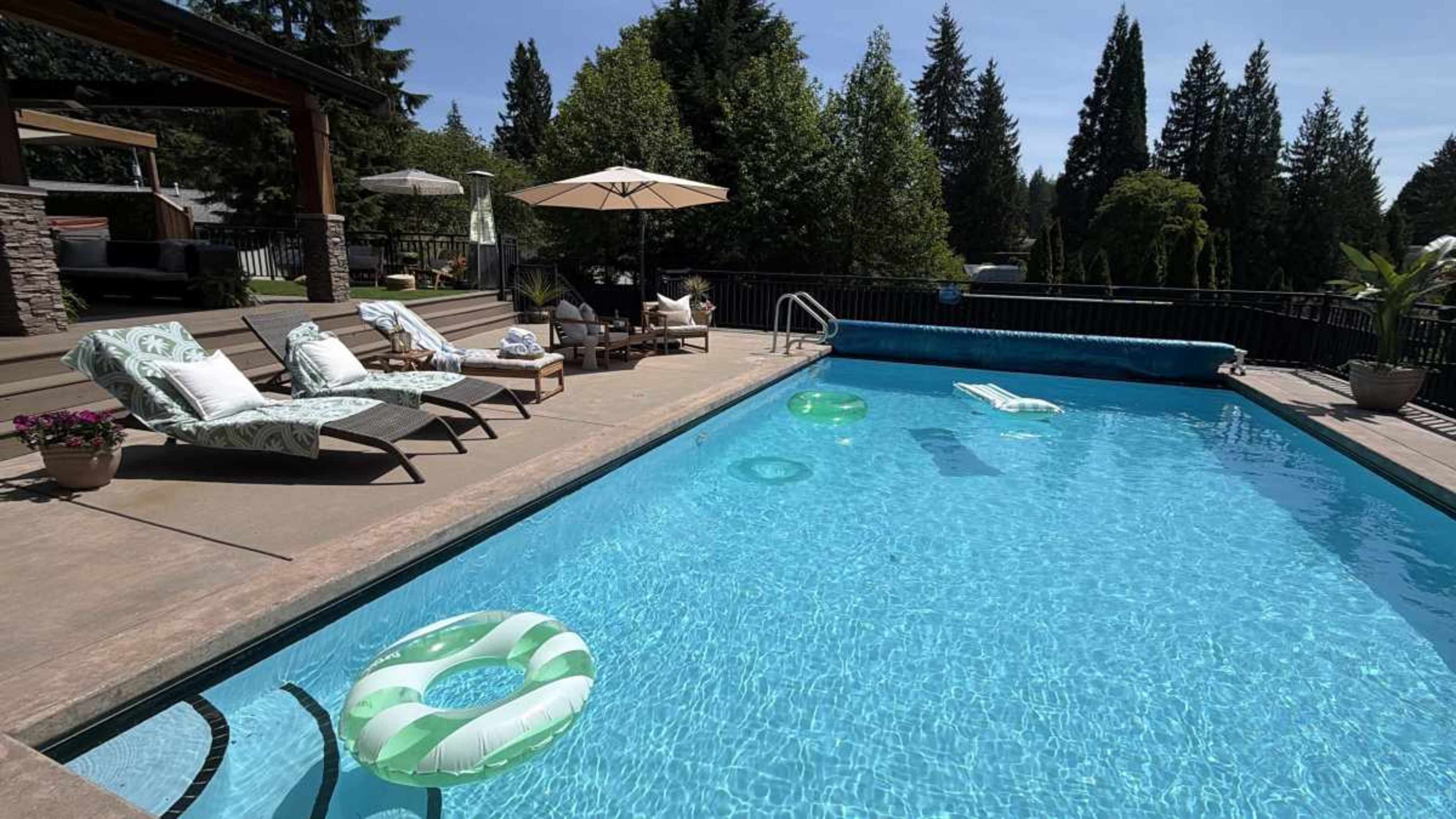 The image shows a swimming pool with clear blue water, surrounded by lounge chairs and green trees in the background.