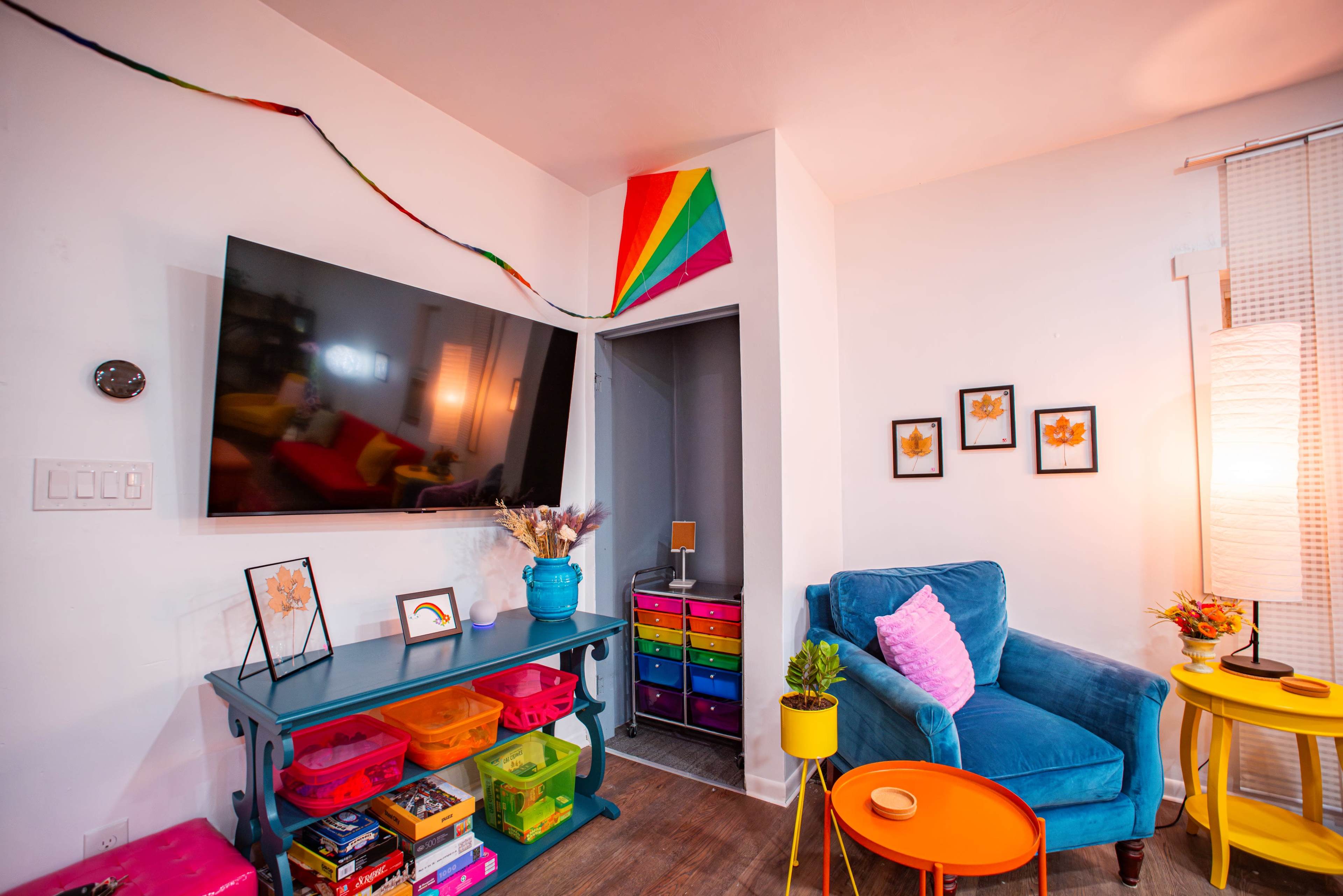 A brightly colored living room features a blue velvet chair, a television mounted on the wall, and shelves filled with vibrant storage bins.