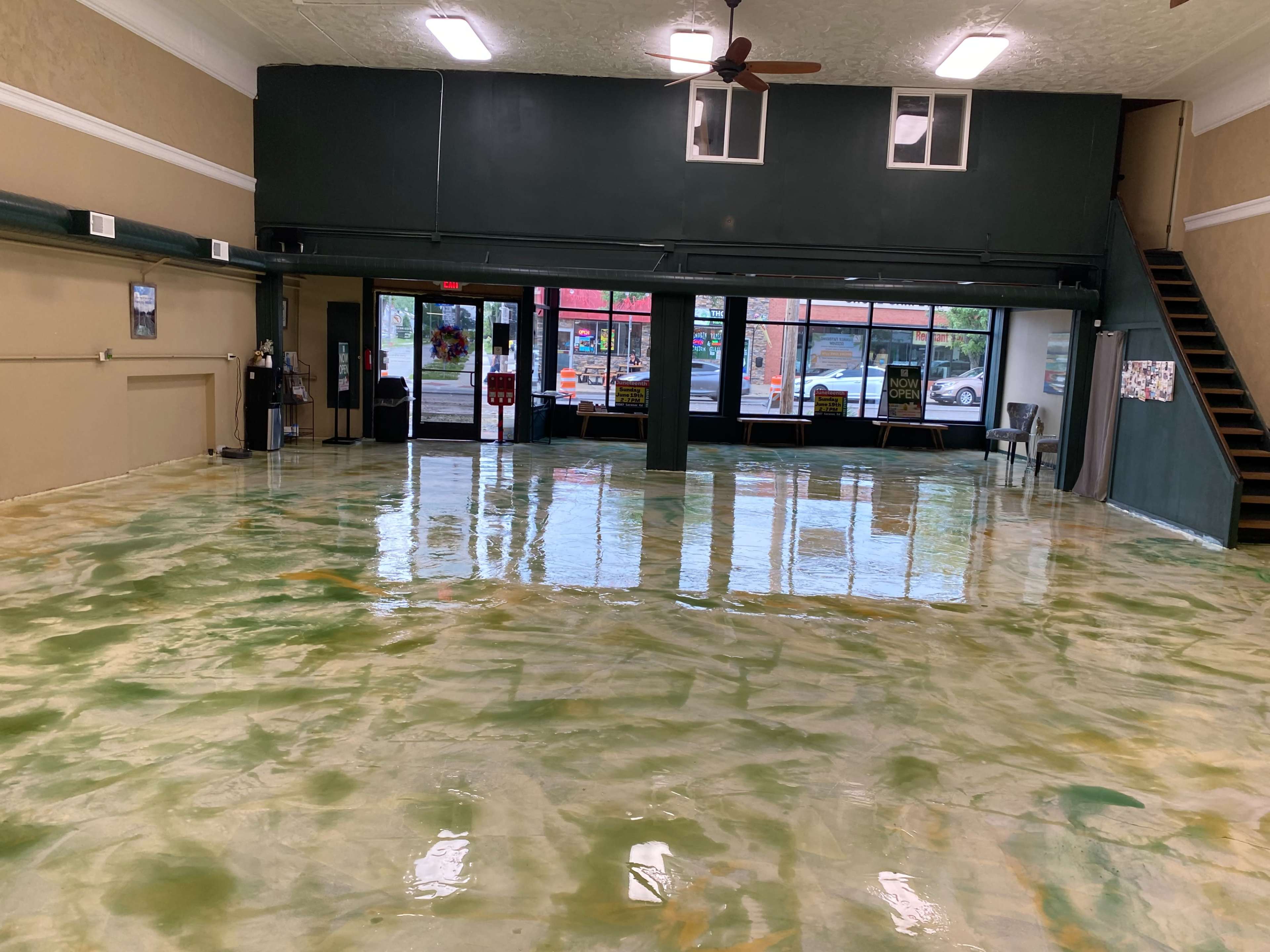 An empty room with a glossy green and beige floor, large windows in the front letting in natural light, and a staircase on the right side.