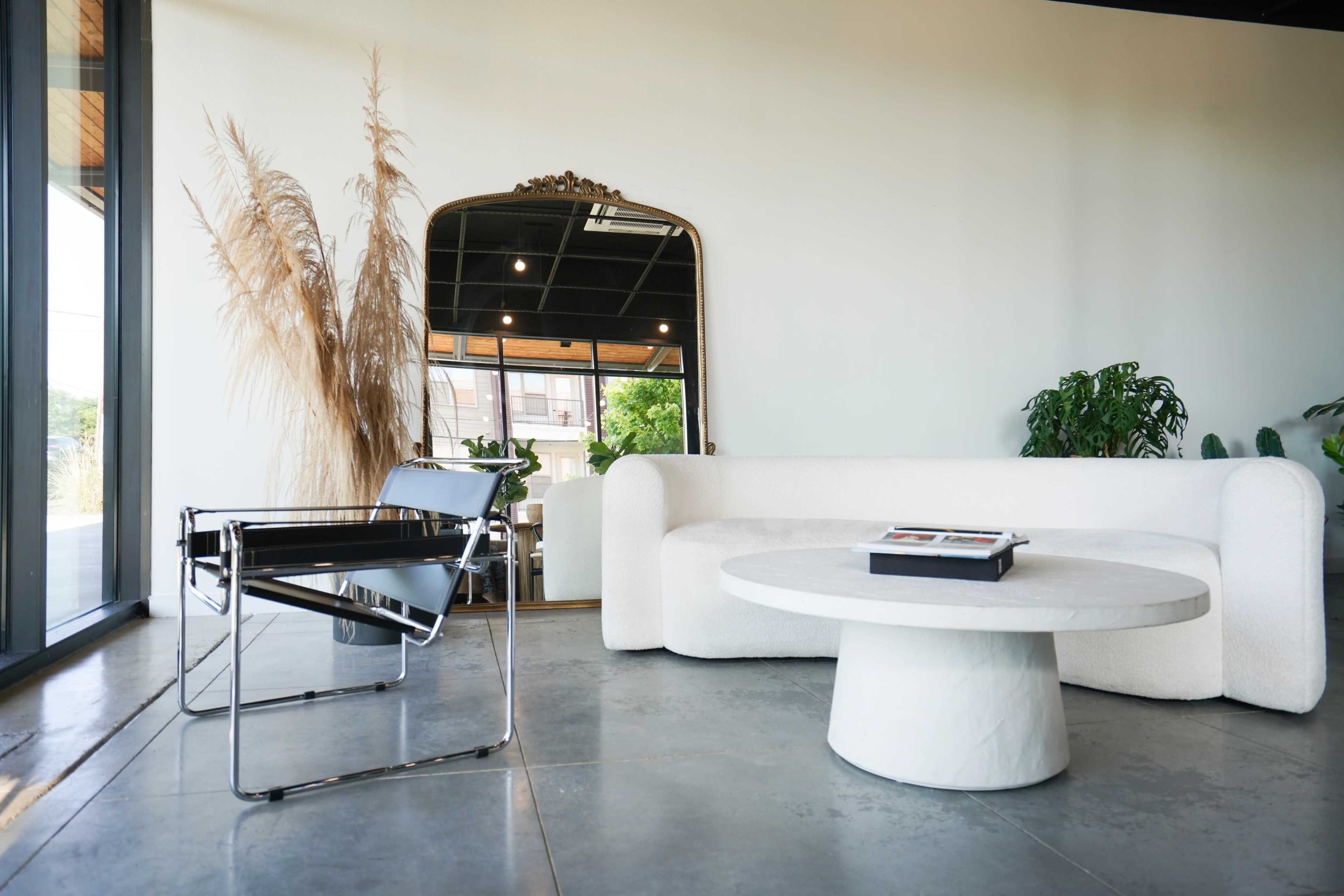 A modern interior featuring a white sofa, a round marble coffee table, and a large mirror beside a tall plant.