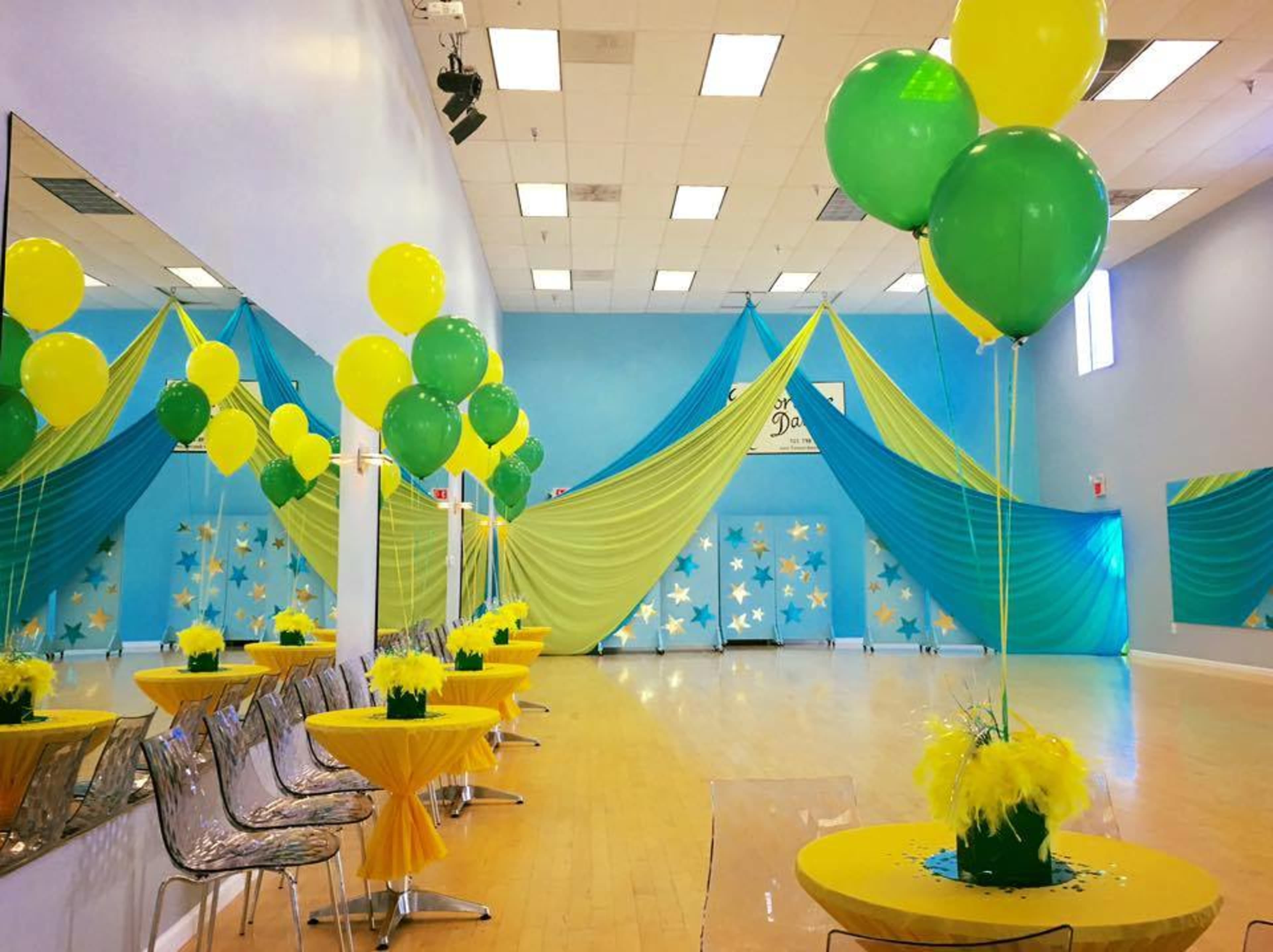 The event space features yellow and green balloons, decorated tables, and draped fabric in blue and teal, creating a vibrant atmosphere.