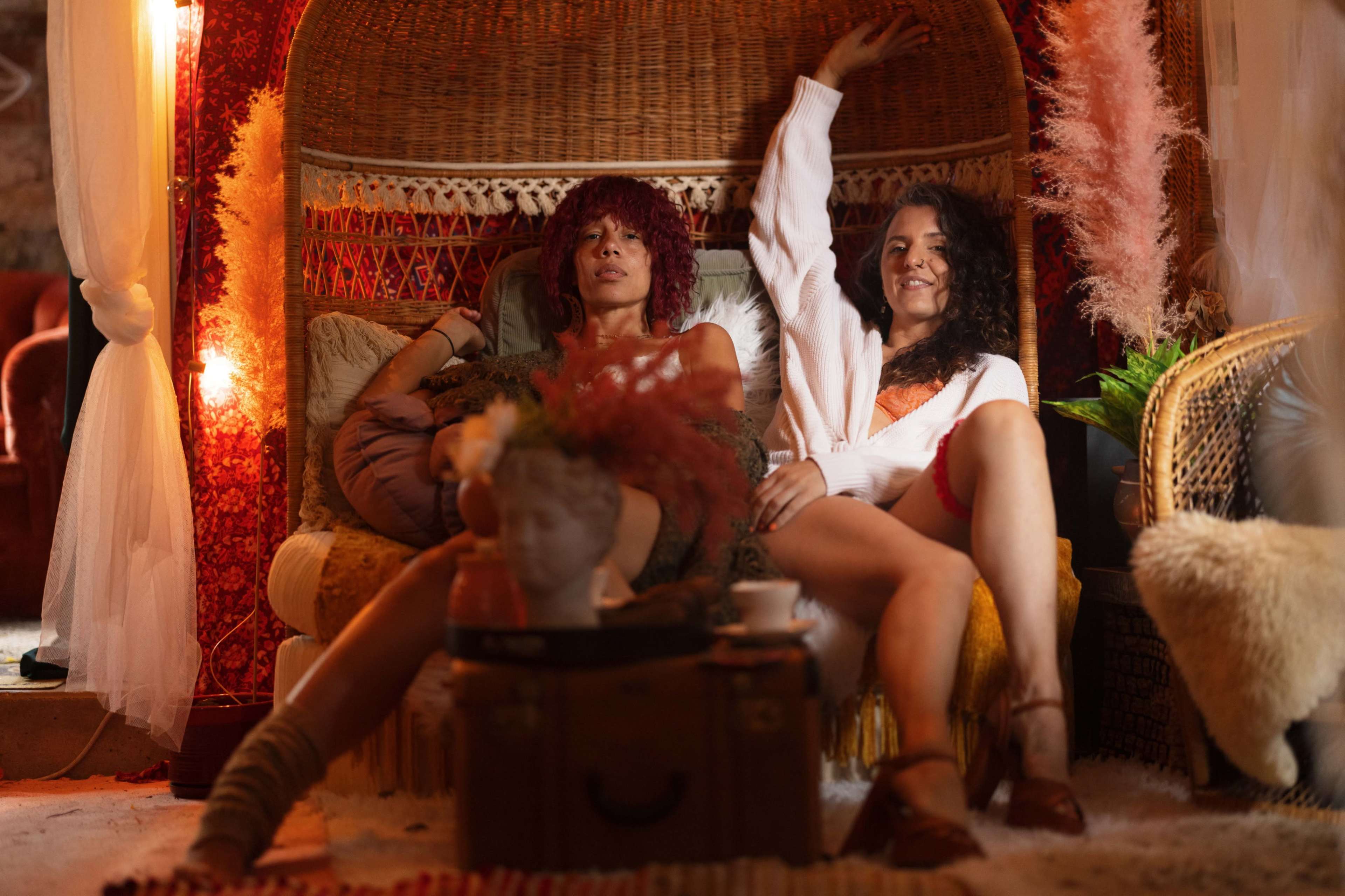Two women sit on a large, woven chair in a cozy room decorated with plants, soft textiles, and warm lighting.