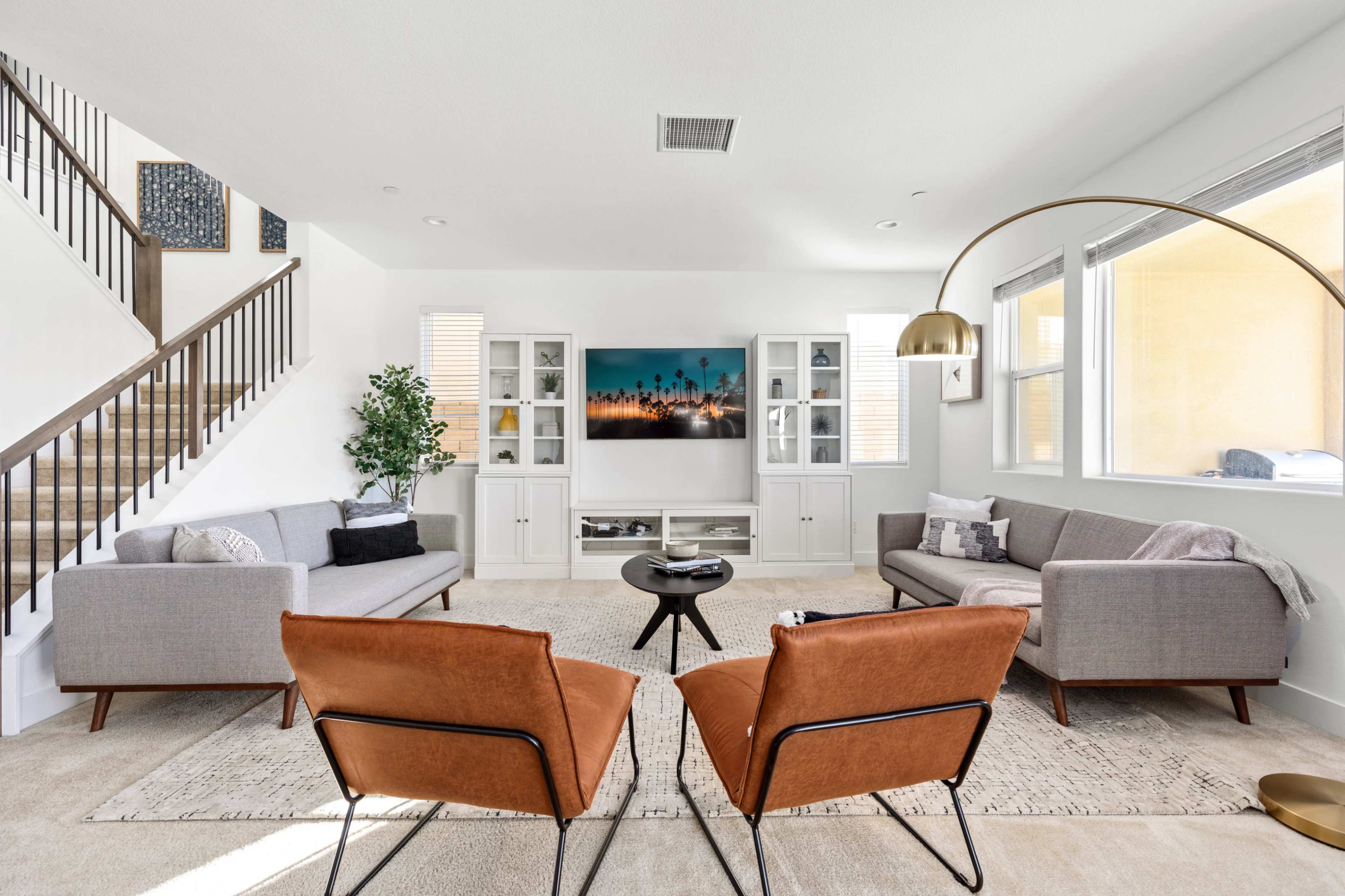 A modern living room features two brown armchairs facing a circular black coffee table, flanked by a gray sofa and a second gray couch, with a television mounted on the wall.