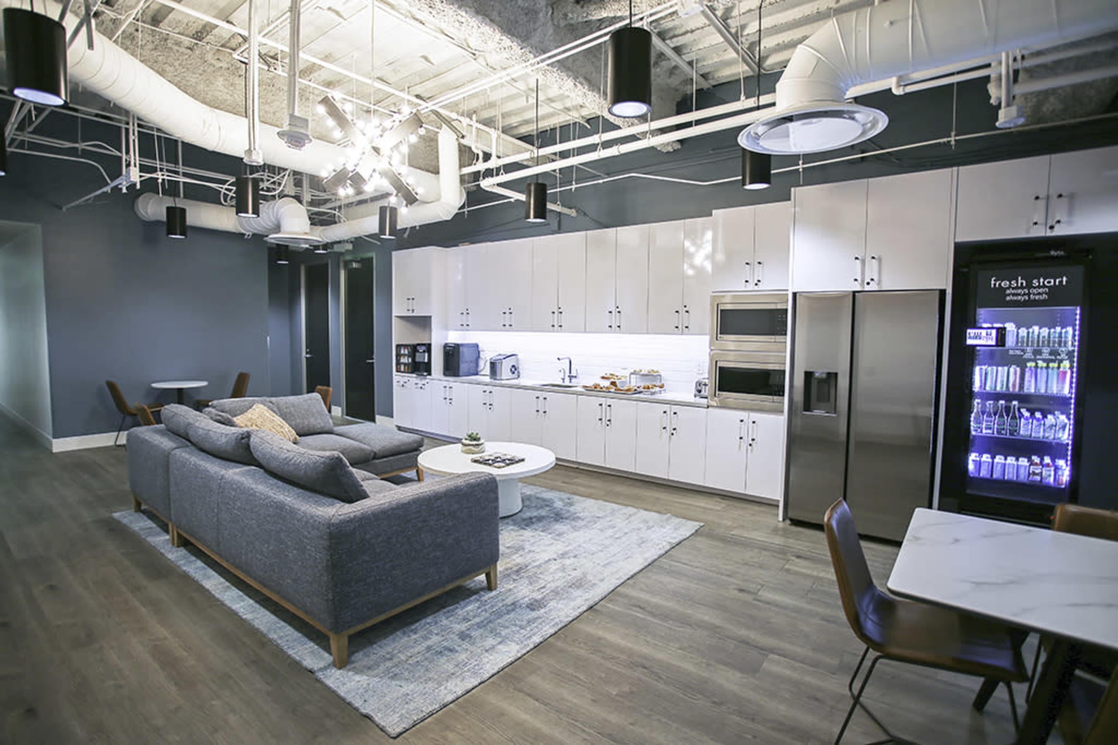 The image shows a modern kitchen and lounge area featuring a grey sofa, a round coffee table, a kitchenette with white cabinets, and a refrigerator stocked with beverages.