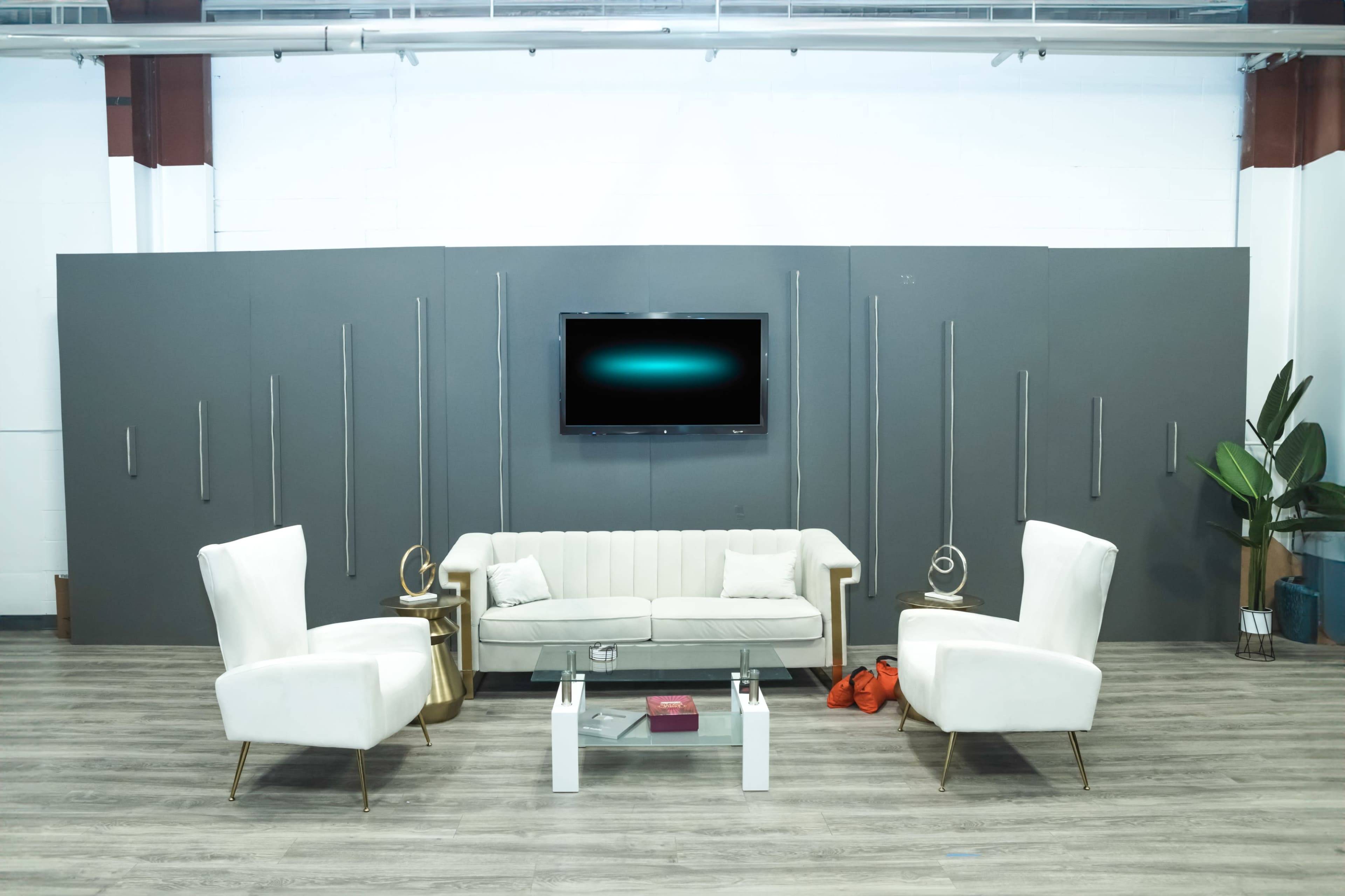 The scene features a modern seating arrangement with a light-colored sofa and two matching armchairs in front of a mounted television, surrounded by a gray wall and a green plant.