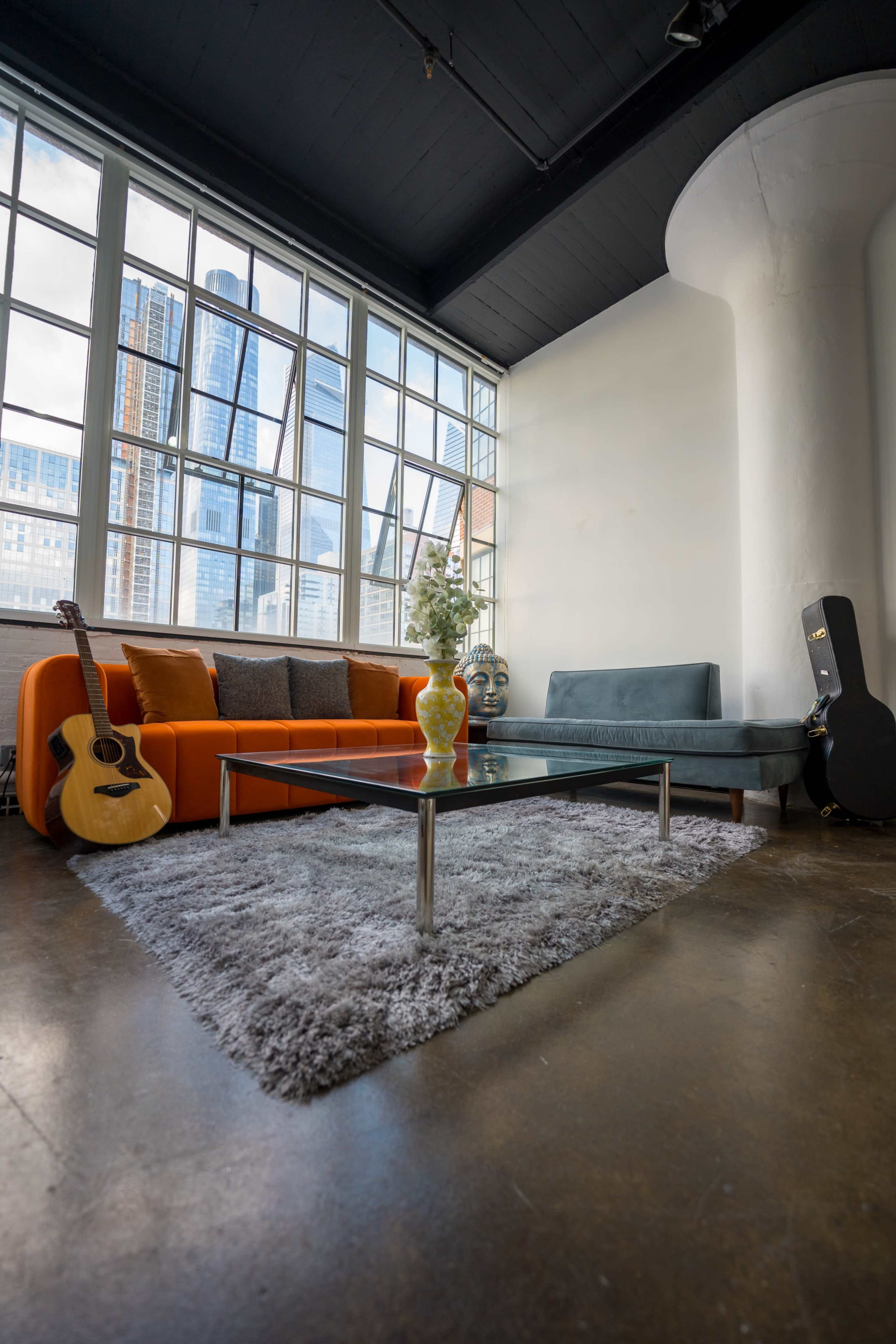 A modern living room with an orange couch, a gray sofa, a glass coffee table, and large windows overlooking a city skyline.