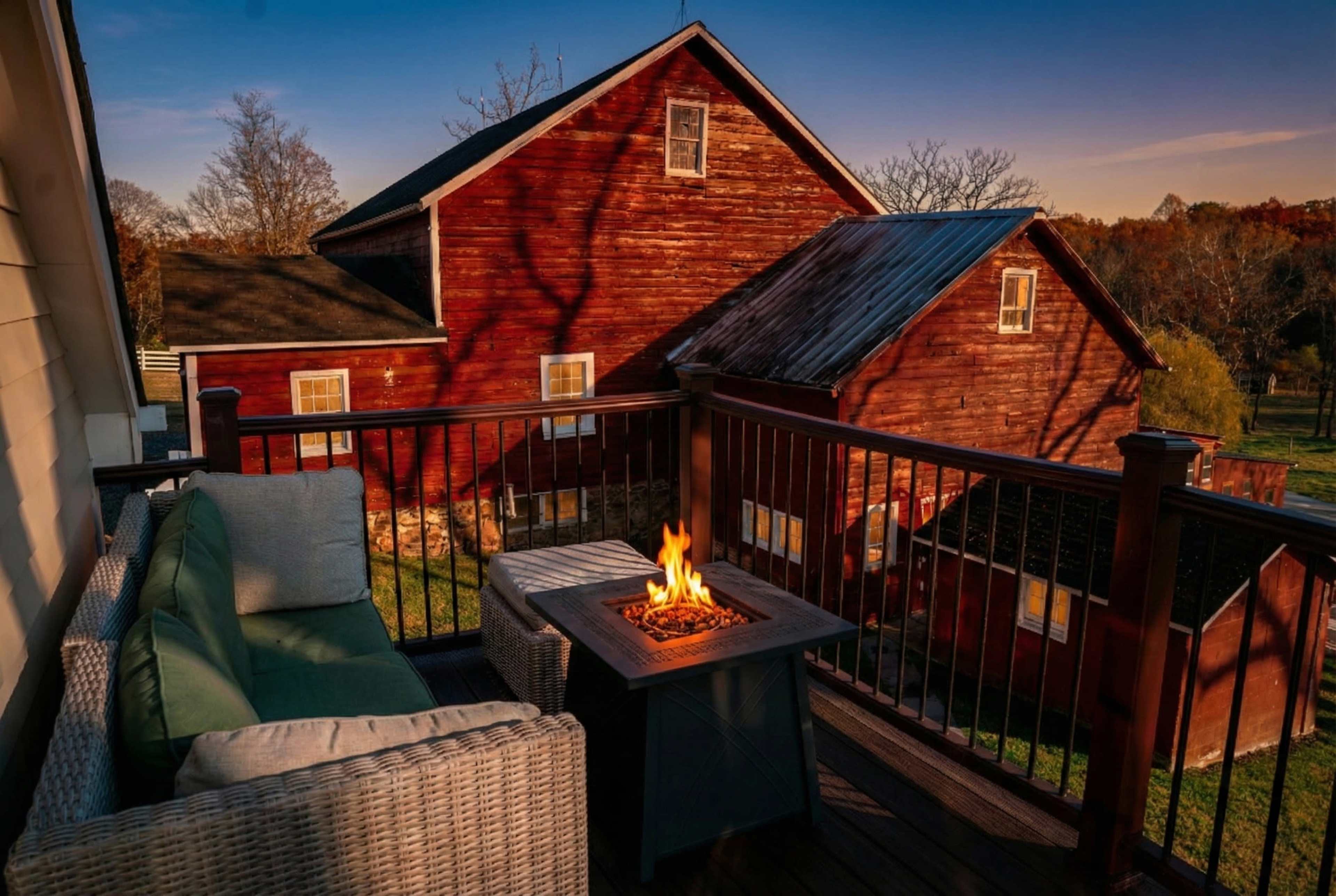 The scene shows a cozy patio with a green sofa and a fire pit, overlooking a rustic red barn and autumn foliage under a clear sky.