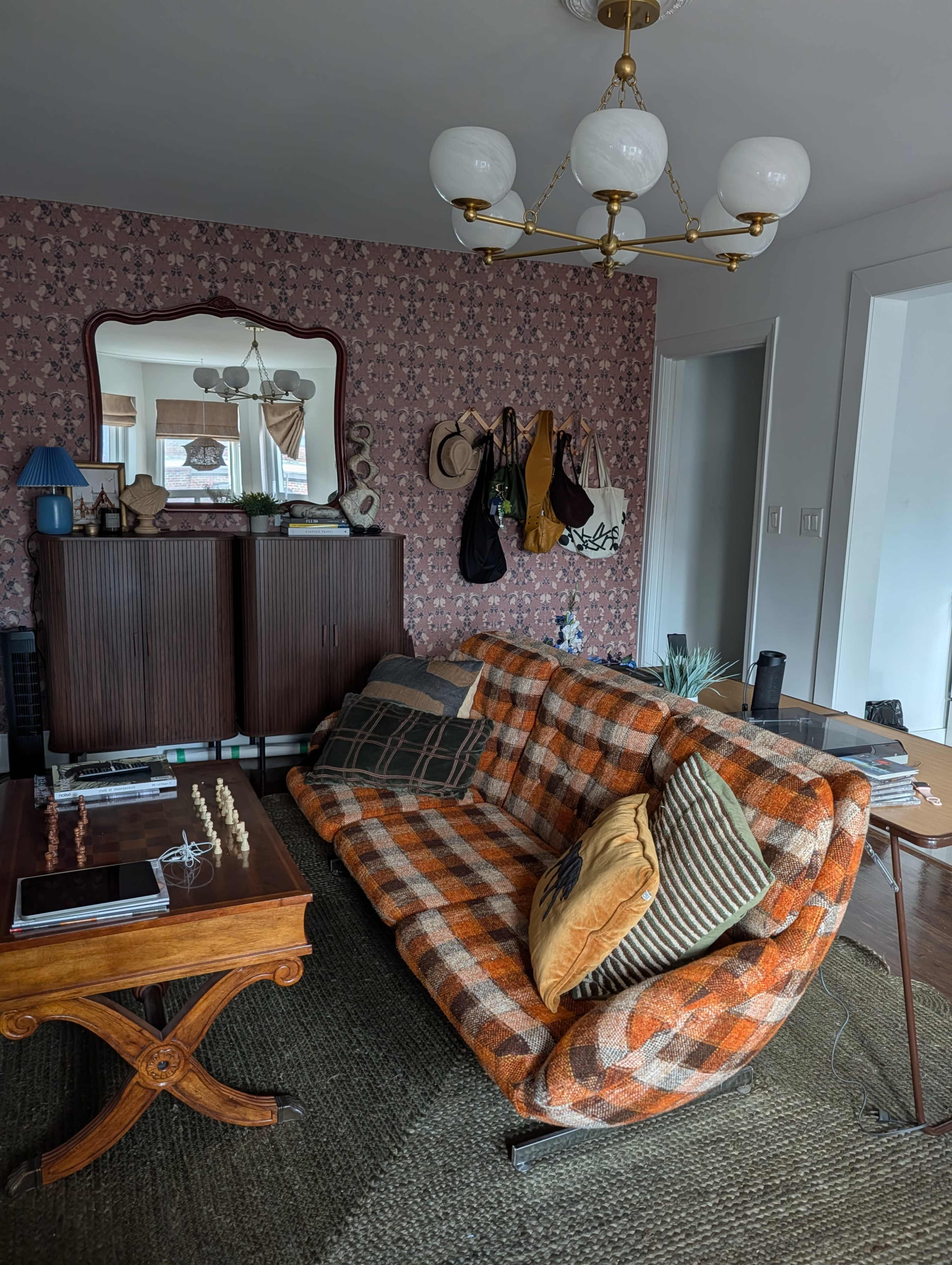The image shows a living room featuring a patterned orange and brown sofa, a wooden table, and various decorative items, with a floral wallpaper background.