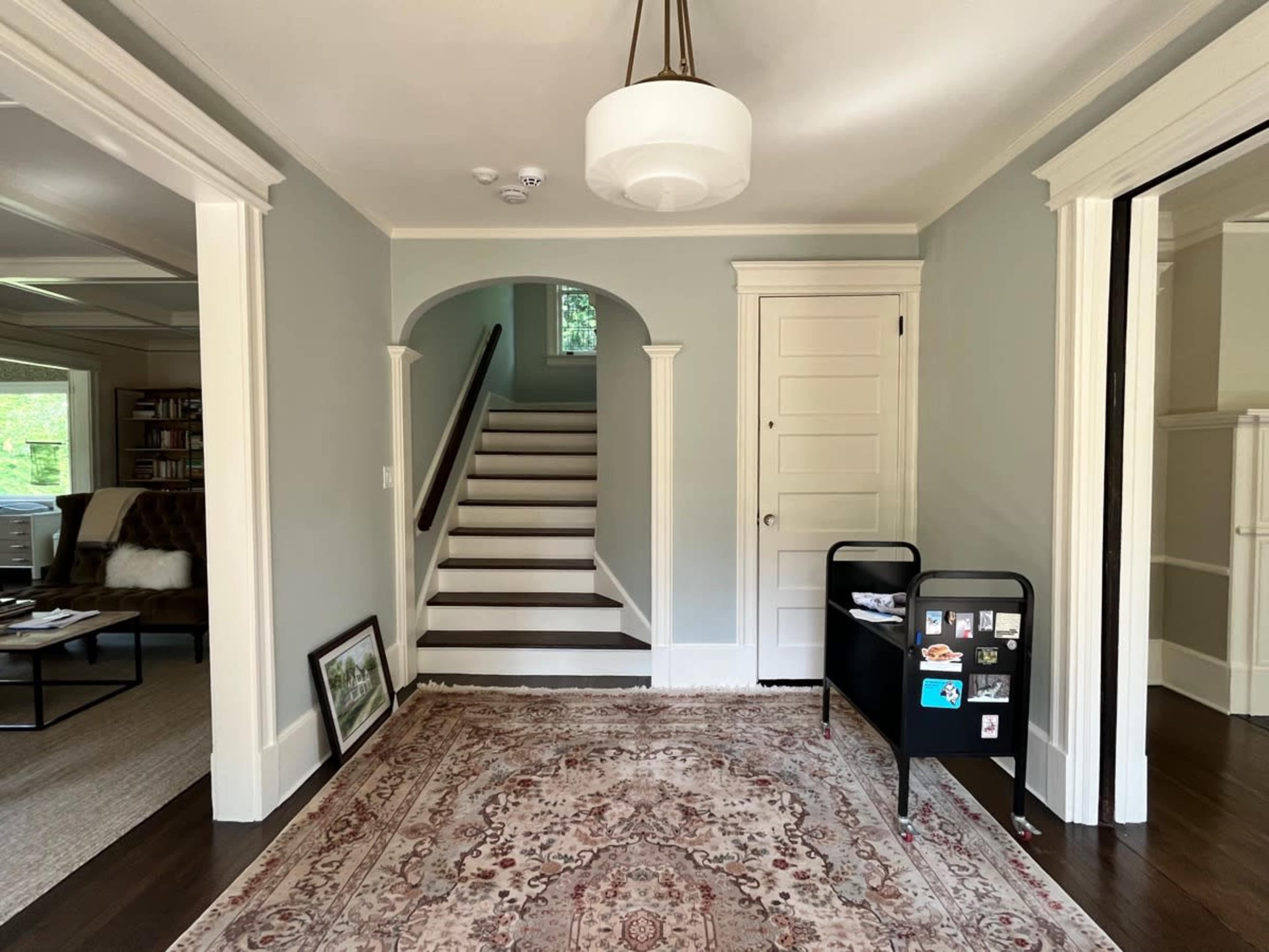 A foyer with a staircase in the background, a decorative area rug on the floor, and a slim black cart displaying various items.