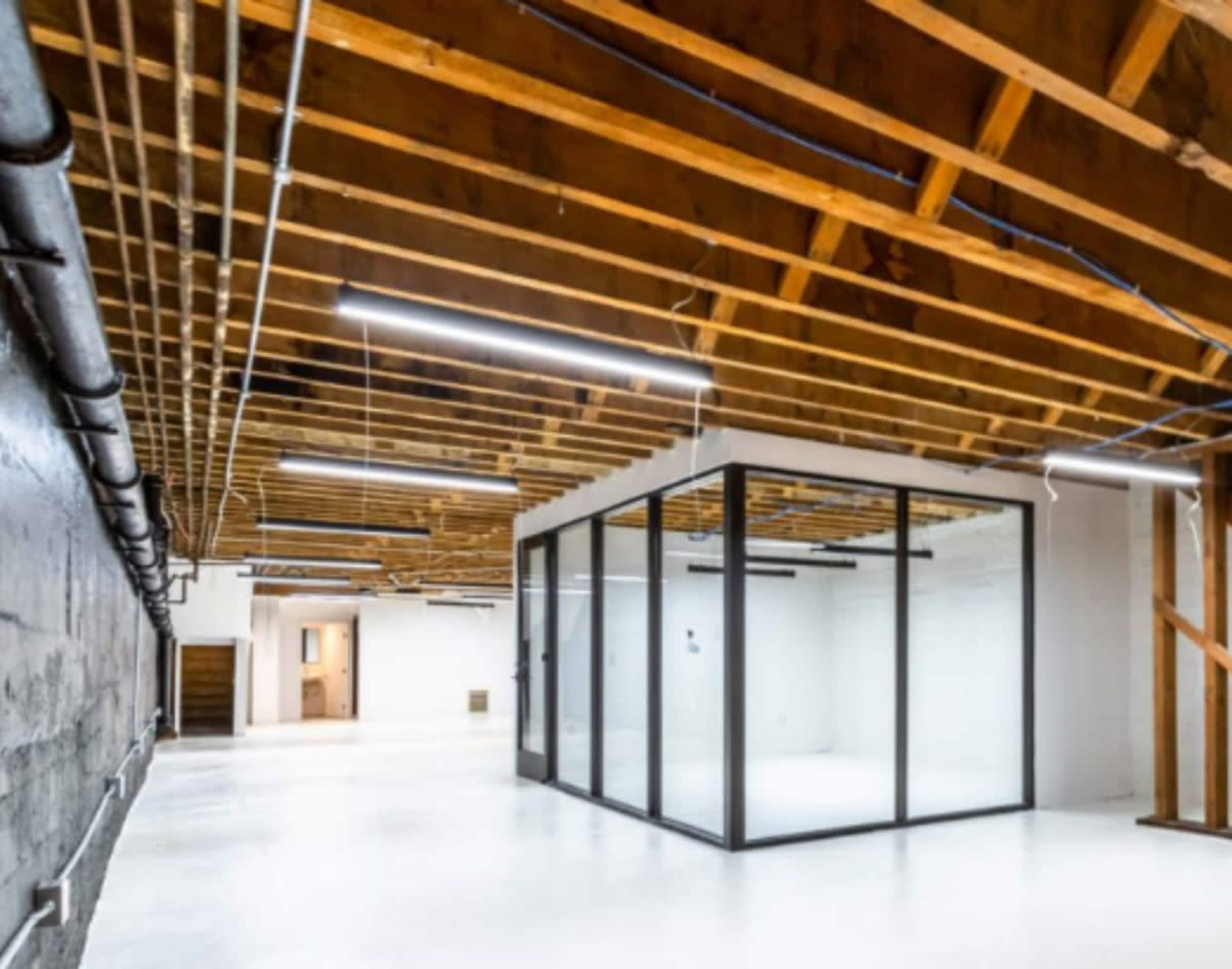 The image shows an empty warehouse-like space with exposed wooden beams, white walls, and a glass-enclosed room in one corner.