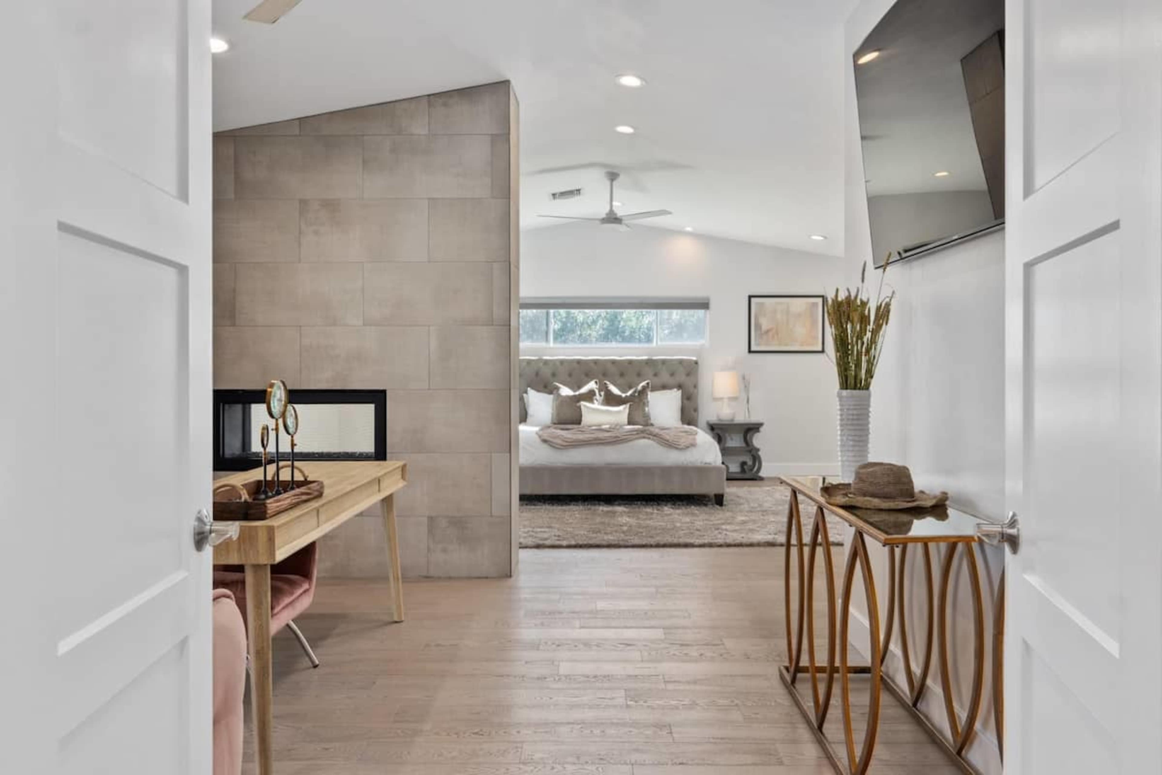 A modern bedroom visible through an open doorway, featuring a bed with decorative pillows, a fireplace, and a minimalist desk with elegant furnishings.