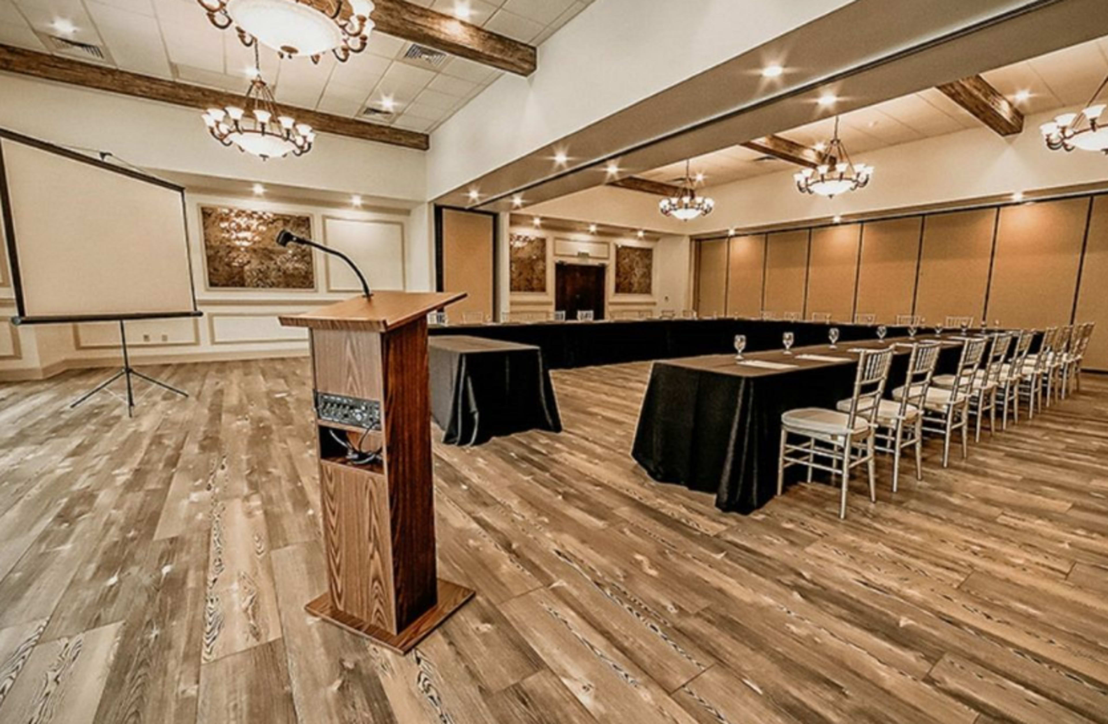 The image shows a conference room with a podium at the front and multiple tables arranged in a U-shape, accompanied by chairs and a projector screen.