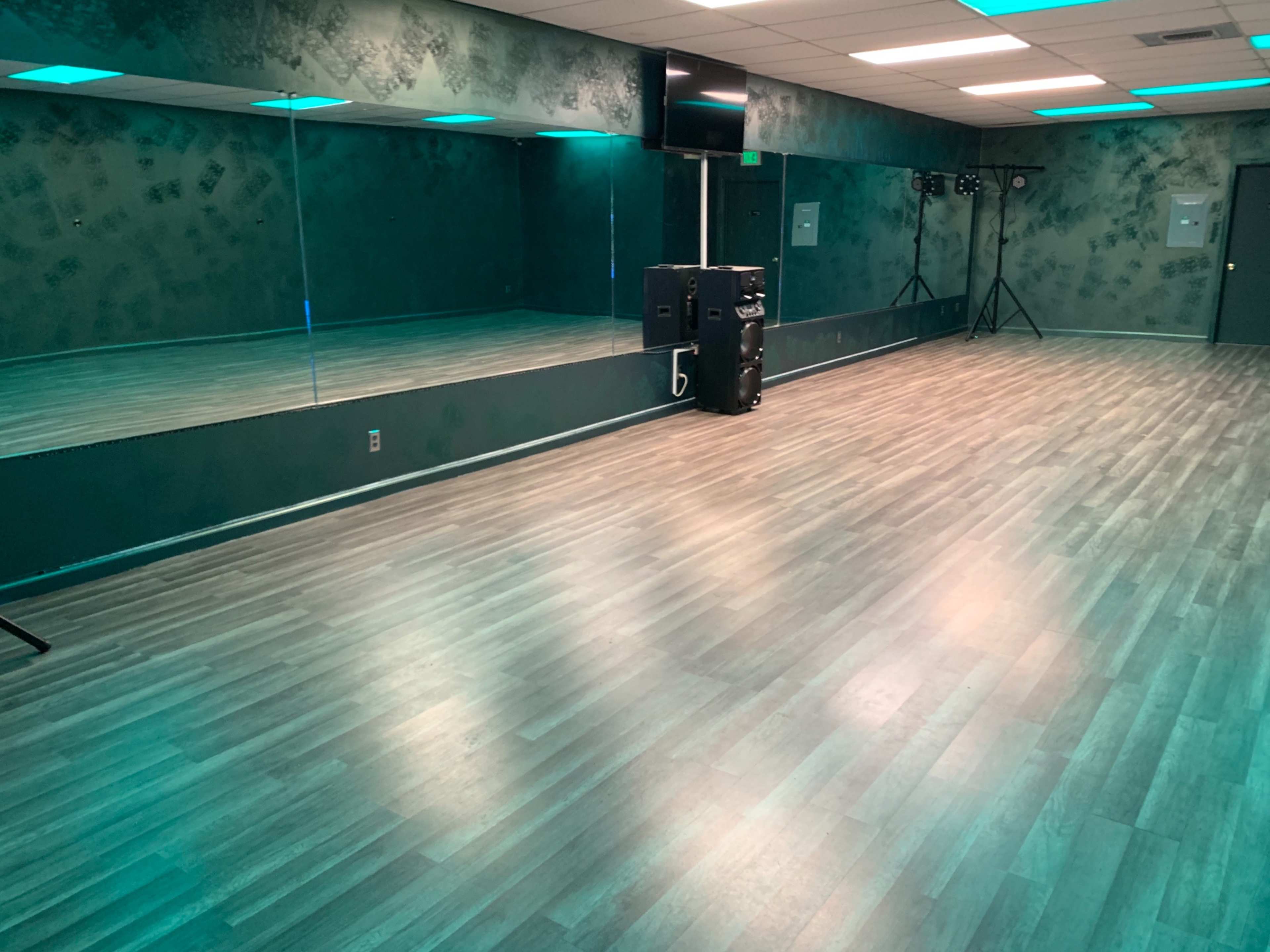 A dance studio with a mirrored wall, wooden flooring, and teal lighting effects.