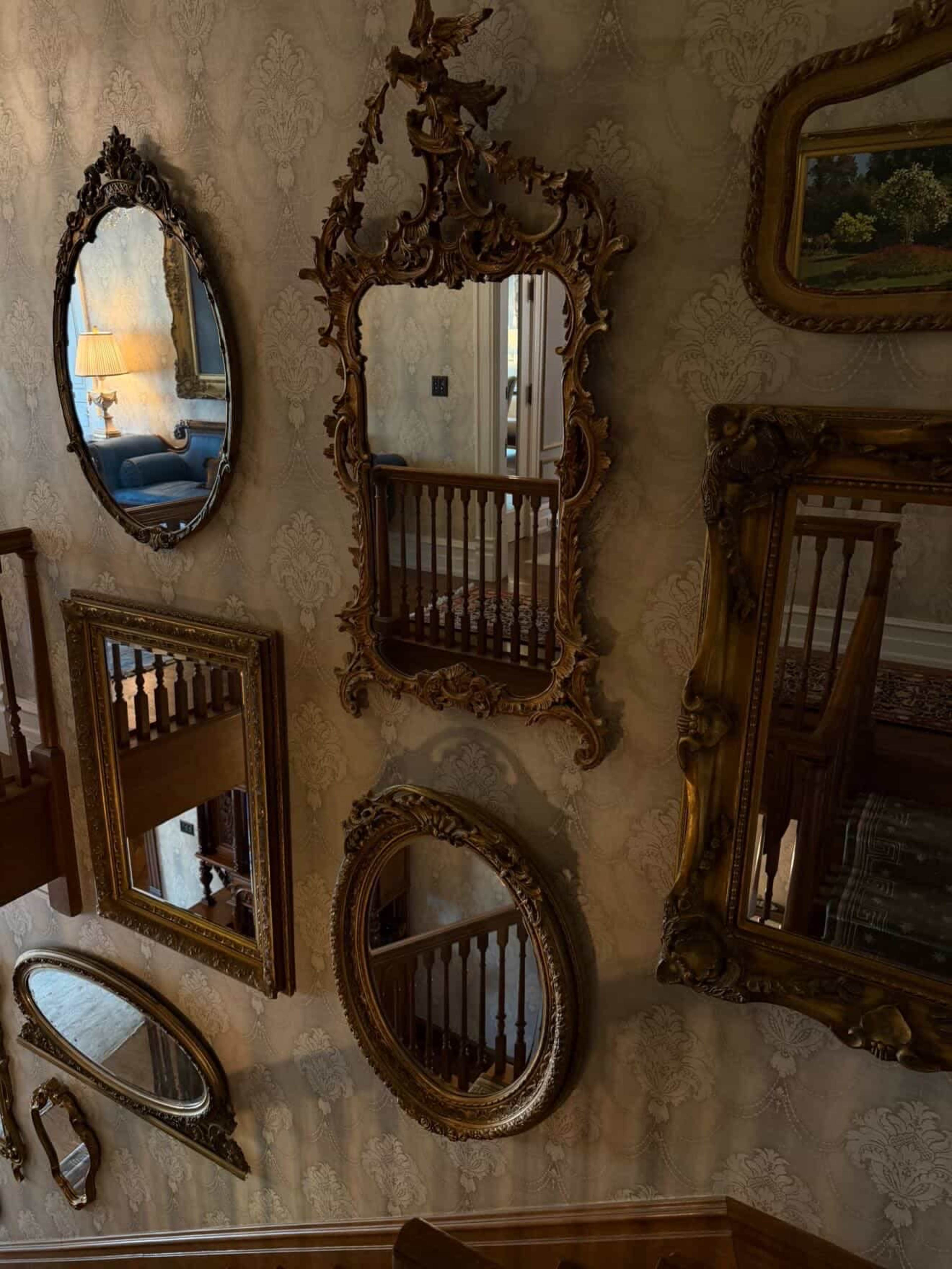 The image shows a wall adorned with several ornate mirrors of various shapes and sizes, reflecting a staircase and a portion of a room in the background.