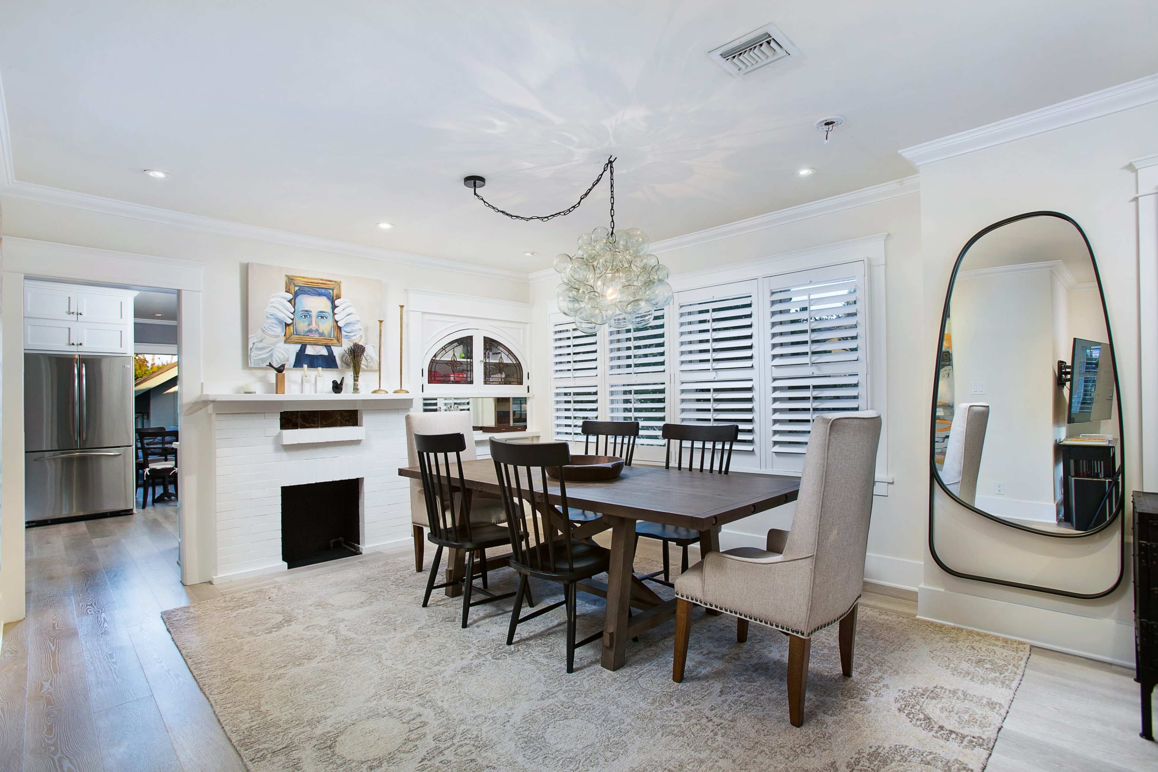 The image features a modern dining room with a wooden table and a mix of black chairs and a beige upholstered chair, a large mirror on the right, and a decorative light fixture above the table.