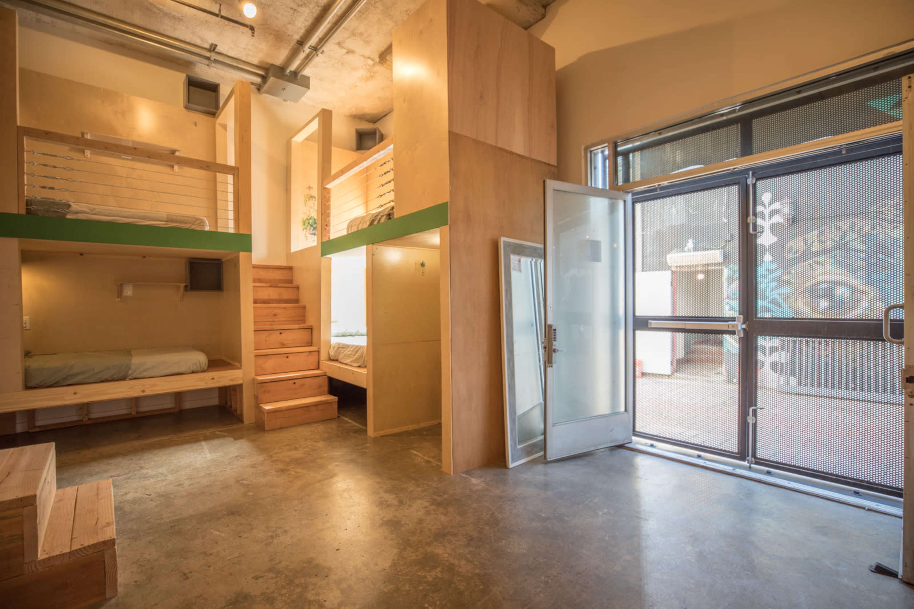 The image shows a modern interior space featuring two elevated sleeping areas, wooden steps leading to one of the beds, and a large glass door that opens to the outside.