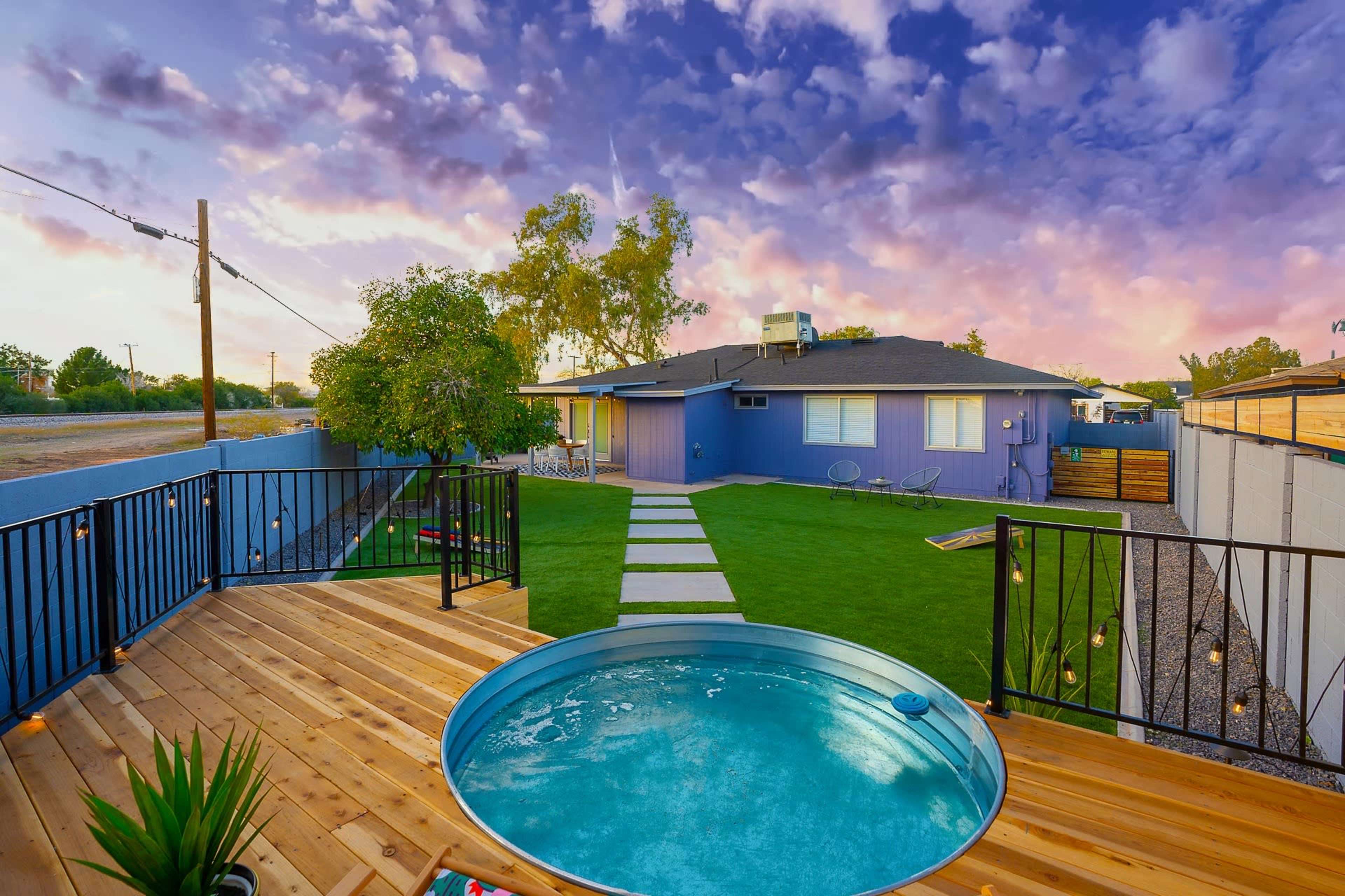 A wooden deck with a circular pool overlooks a grassy yard and a house painted purple, set against a colorful sky.