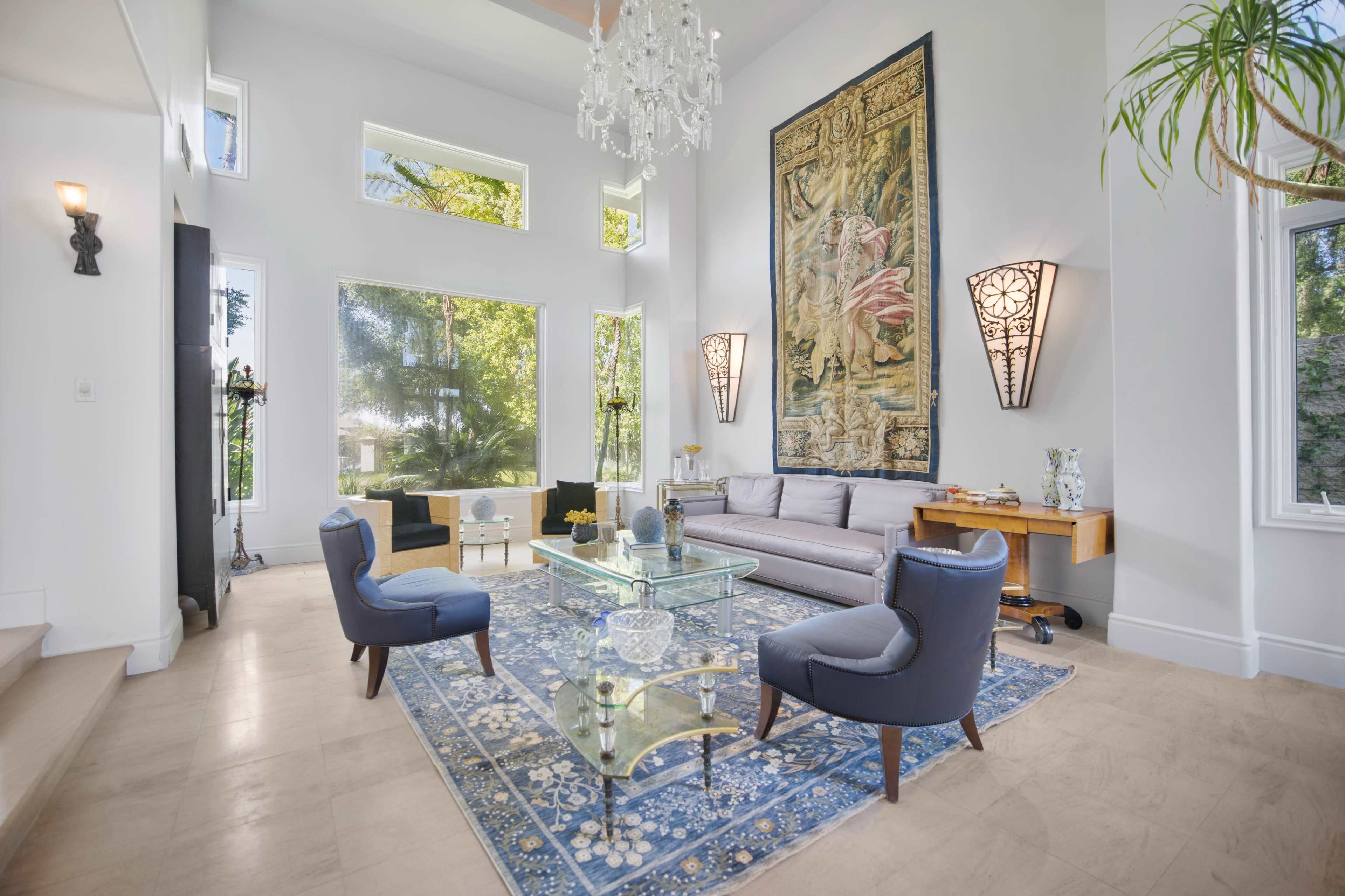 The living room features a chandelier, a large tapestry on the wall, a glass coffee table, and modern furniture arranged around a decorative rug.