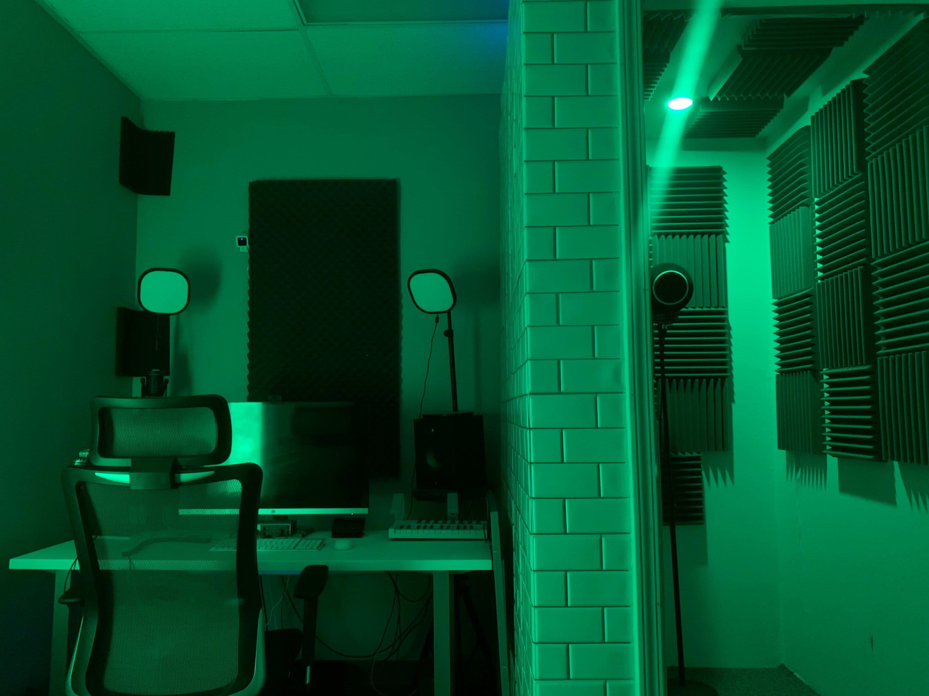 A soundproof recording studio features a desk with a computer and audio equipment, surrounded by acoustic panels and a green ambient light.
