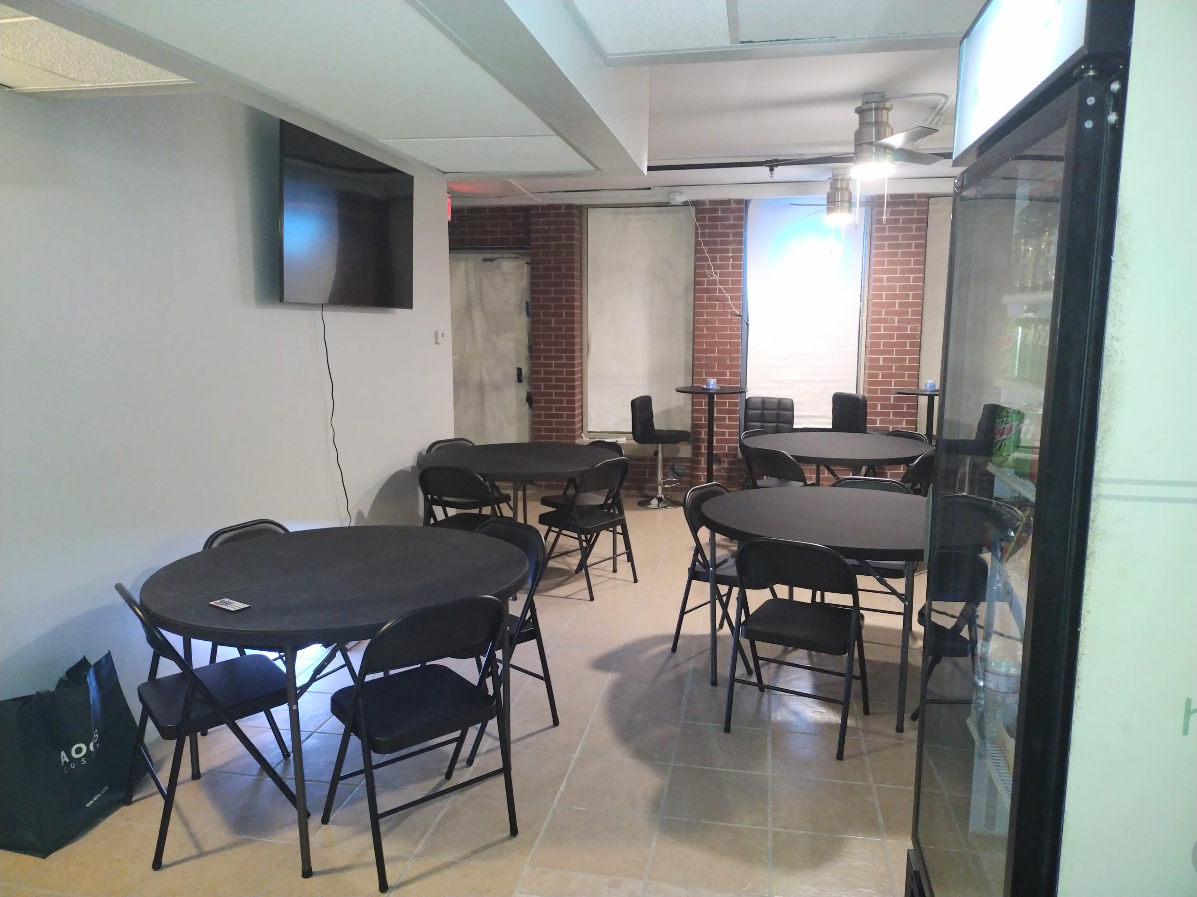 A spacious room with several round tables and black folding chairs, a television on the wall, and a fridge in the corner.