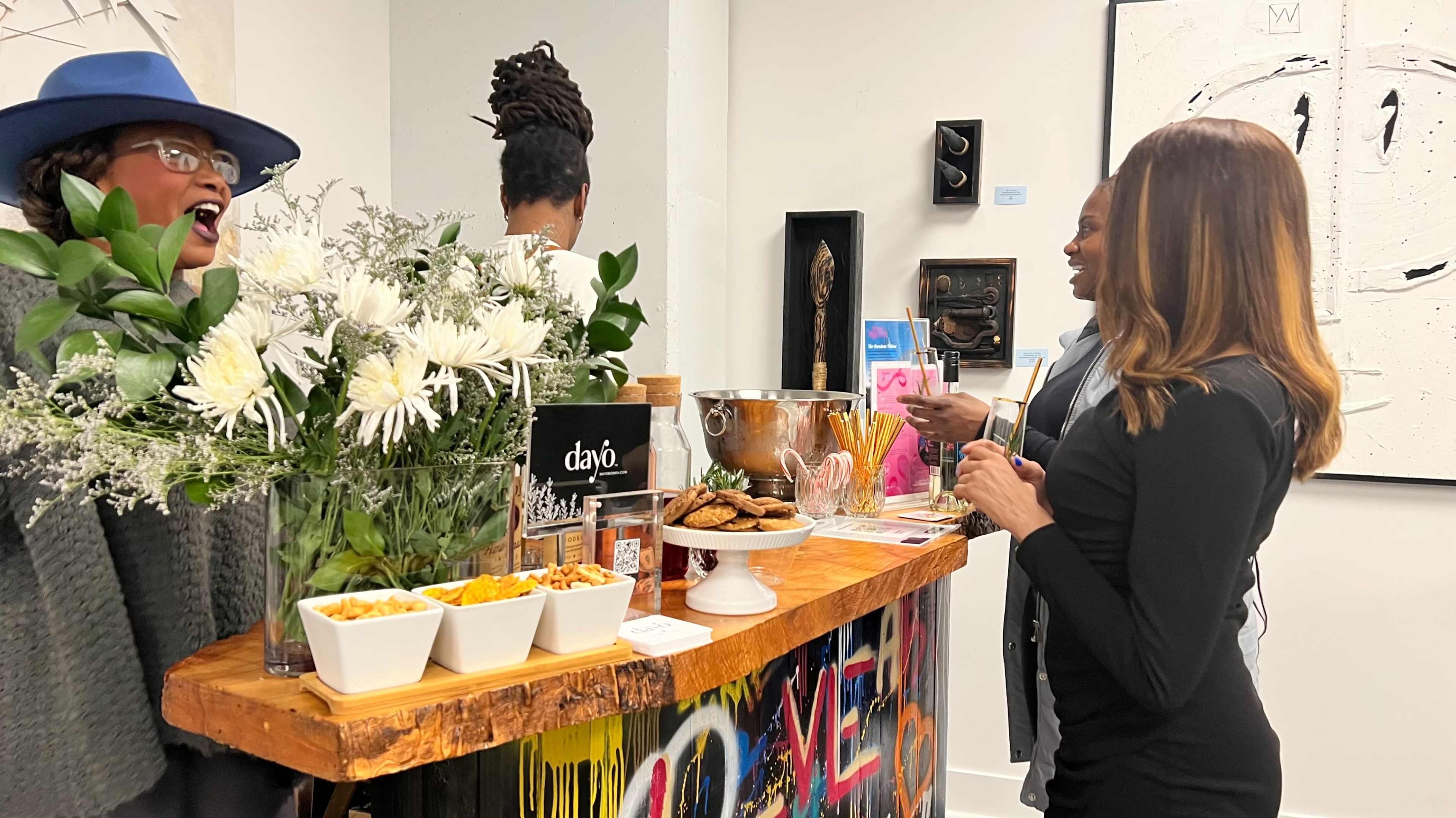 A person in a blue hat laughs while another person talks at a wooden bar adorned with flowers and snacks in a gallery setting.