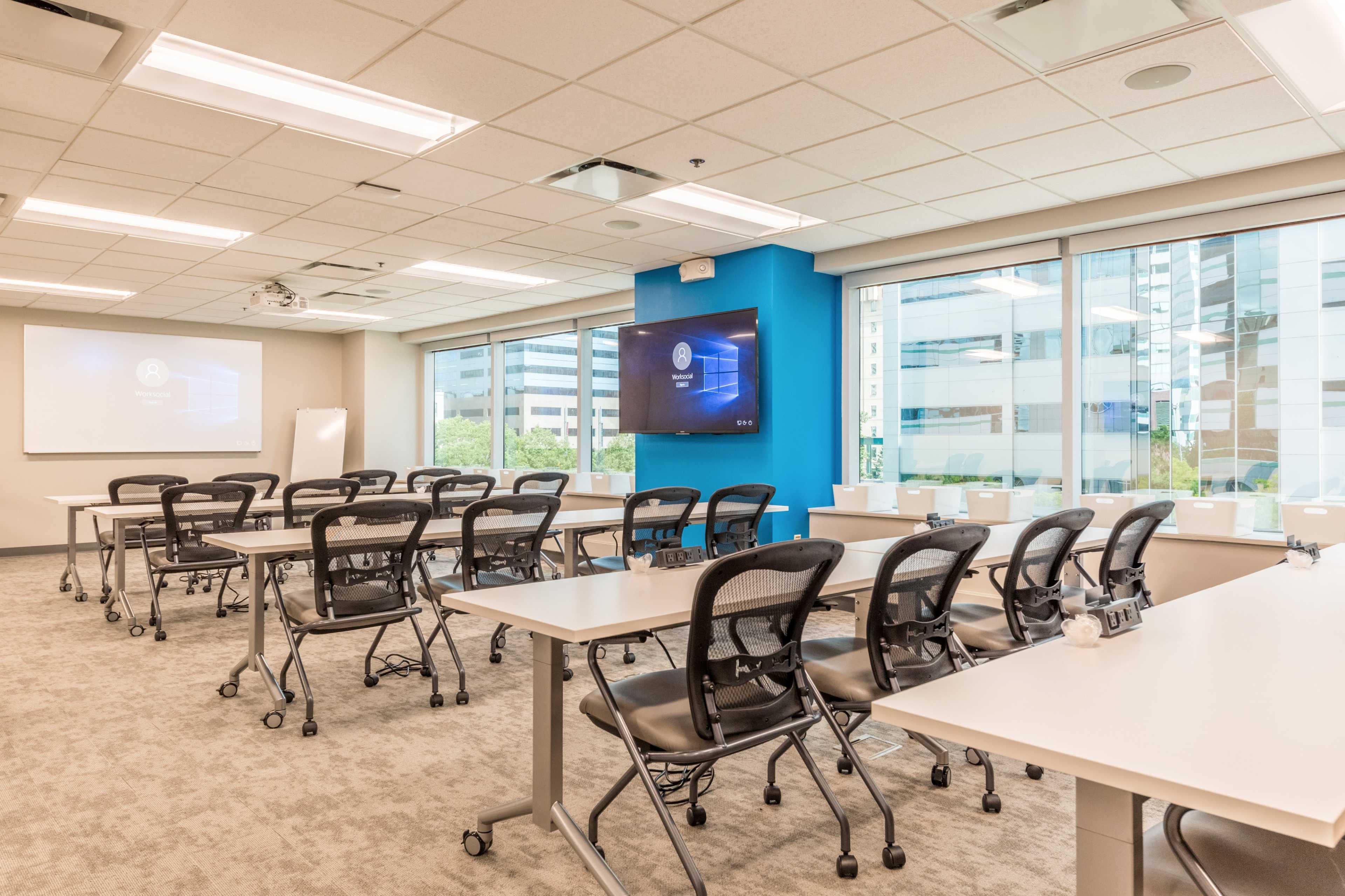 A modern conference room features rows of tables and chairs, with large windows and a wall-mounted screen displaying a presentation.