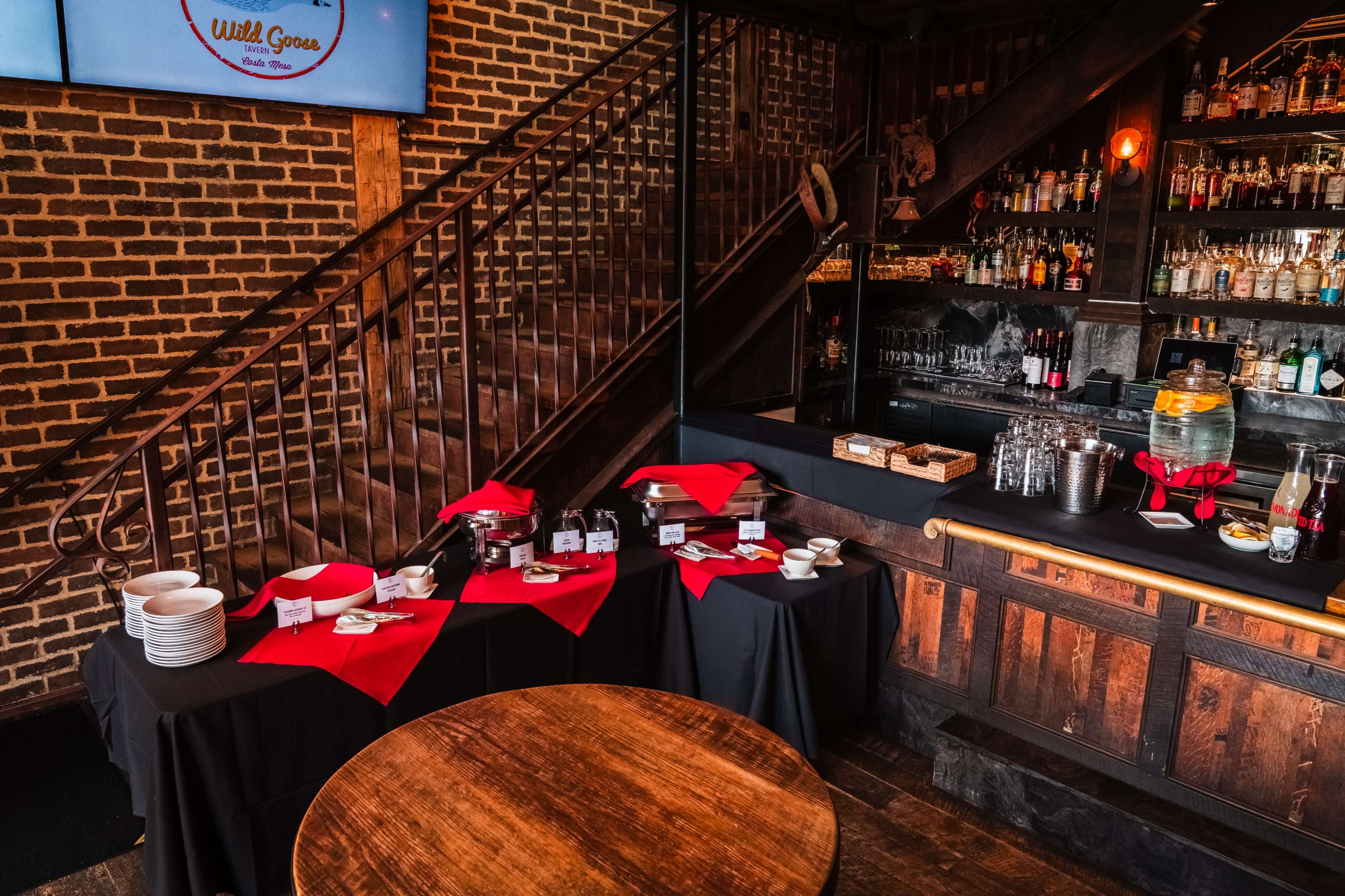 A bar area is set up with a wooden counter, a selection of drinks, glassware, and food options on tables covered with red tablecloths against a backdrop of exposed brick walls and a staircase.