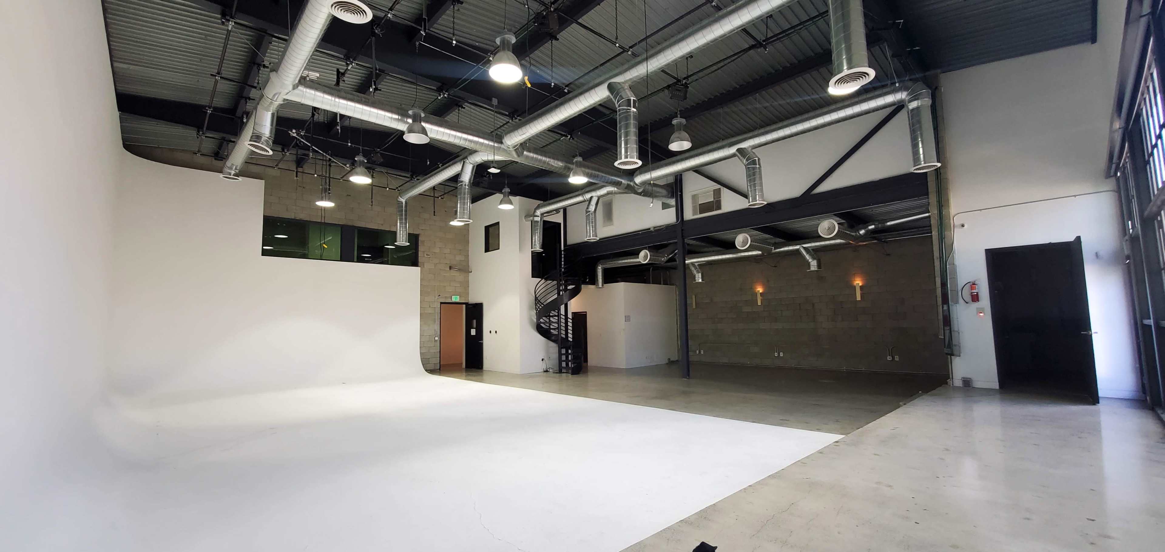 A spacious, empty studio with a large white backdrop, high ceilings, and ventilation ducts visible.
