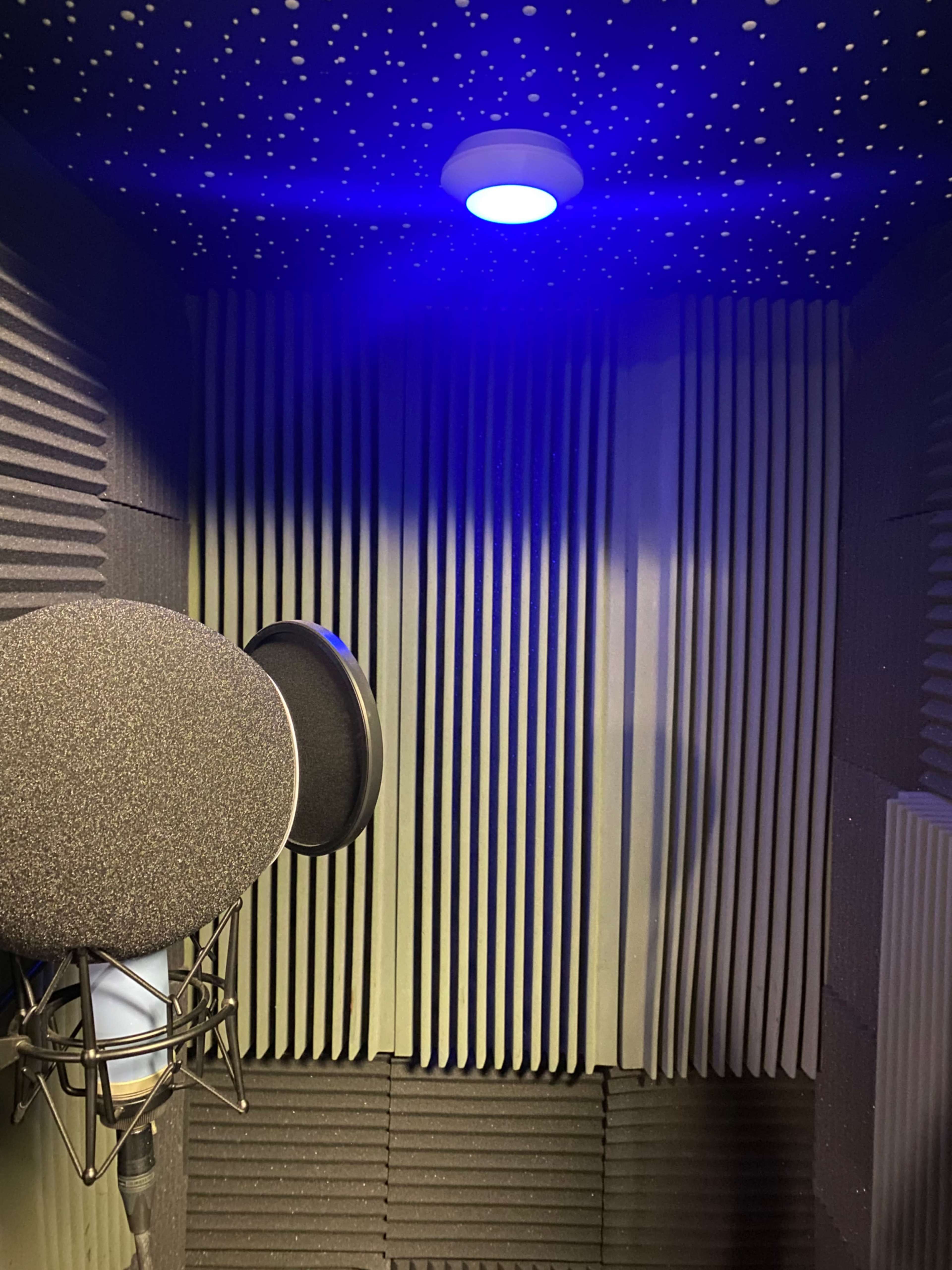 The interior of a soundproof recording booth featuring acoustic foam panels, a microphone with a pop filter, and a ceiling light.